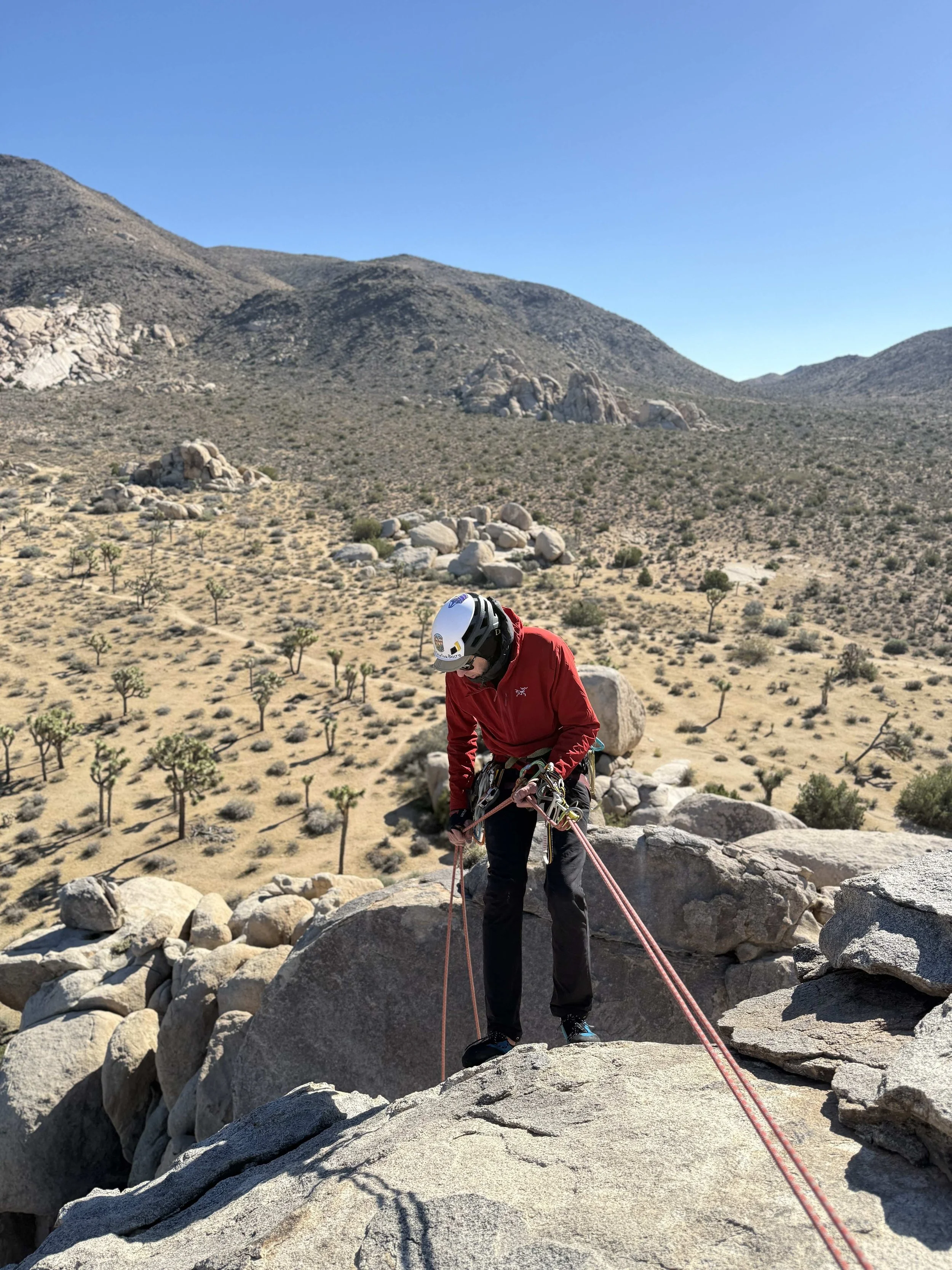 A person in a red jacket and climbing helmet rocks climbing on a large granite boulder in a desert landscape with Joshua trees and rocky mountains in the background.