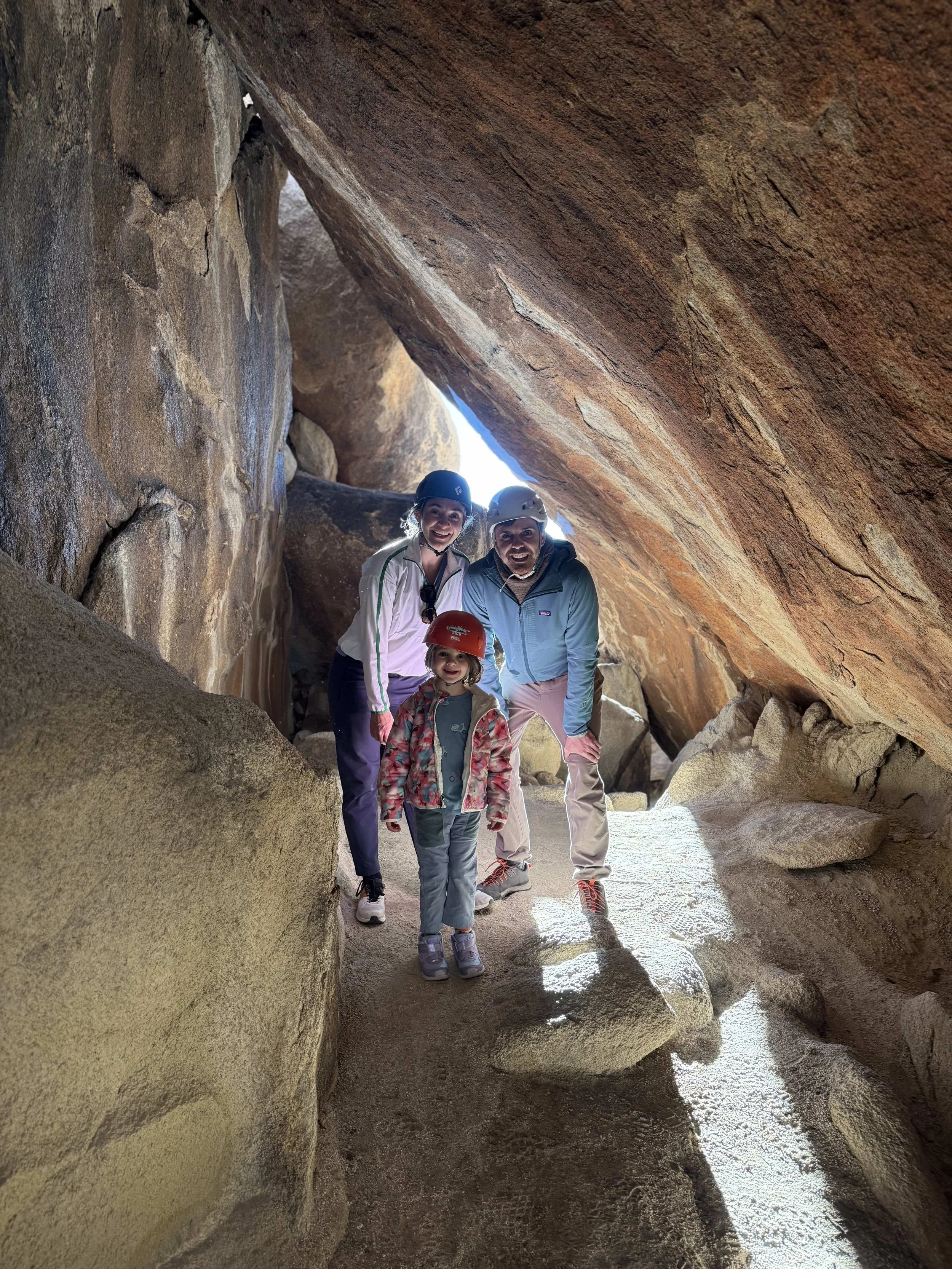 A family of four in helmets exploring a rocky canyon, standing on sand and surrounded by large sandstone walls.