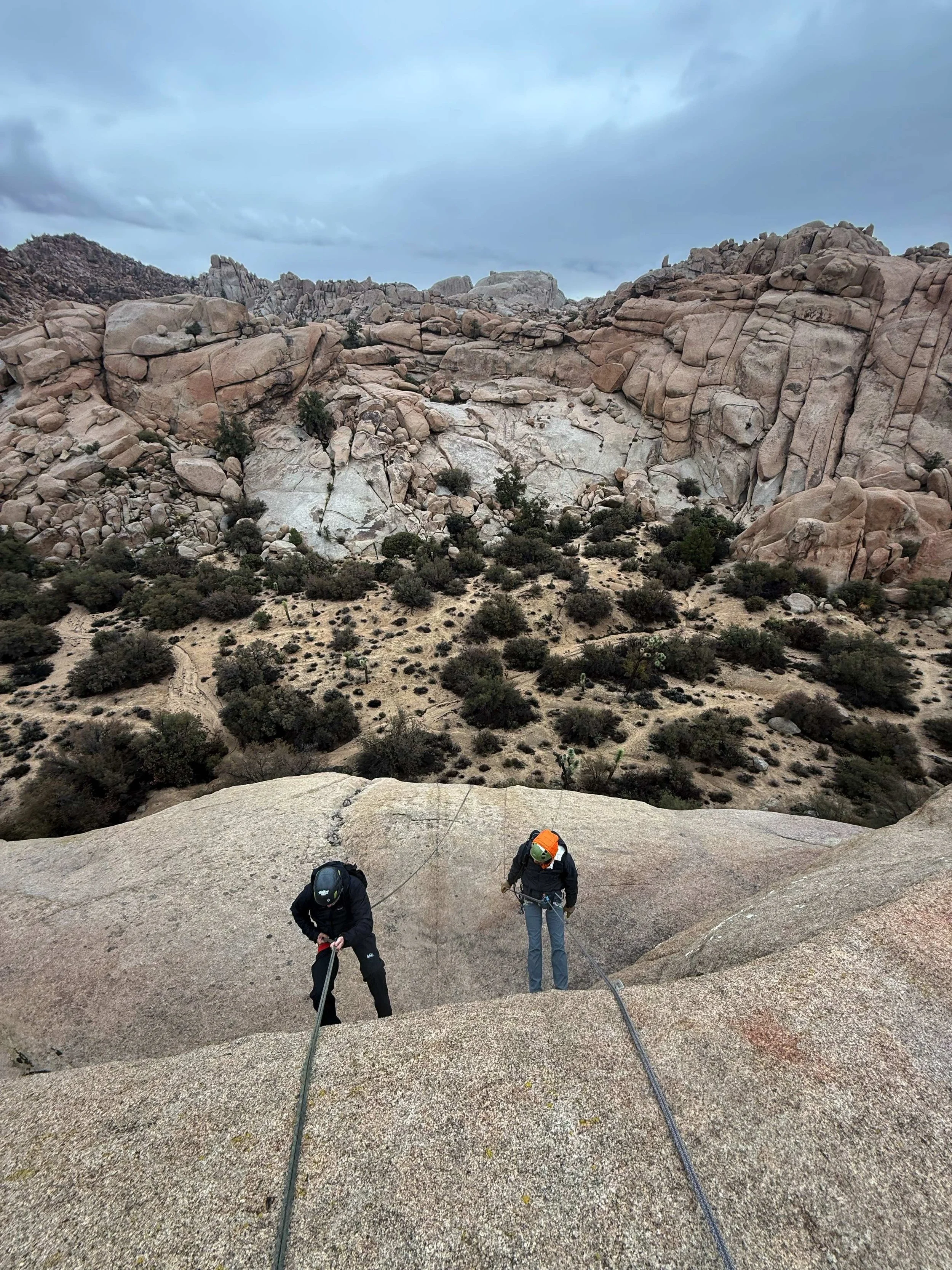 Two climbers ascending a rocky, steep incline in a desert landscape with large beige and pink rocks and sparse vegetation beneath a cloudy sky.