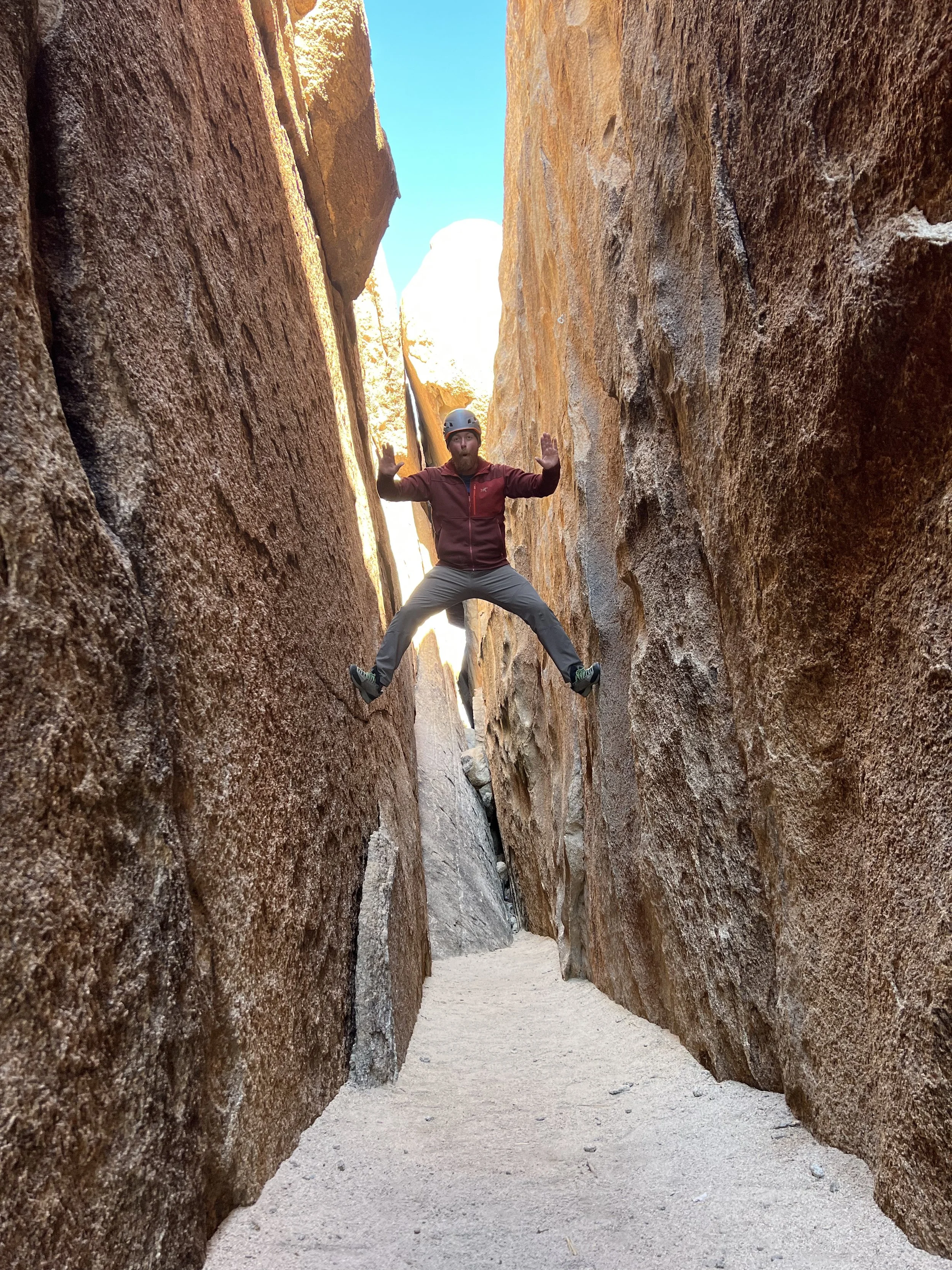 A man in a helmet and outdoor clothing jumps with his legs spread wide and arms raised in a rock canyon.