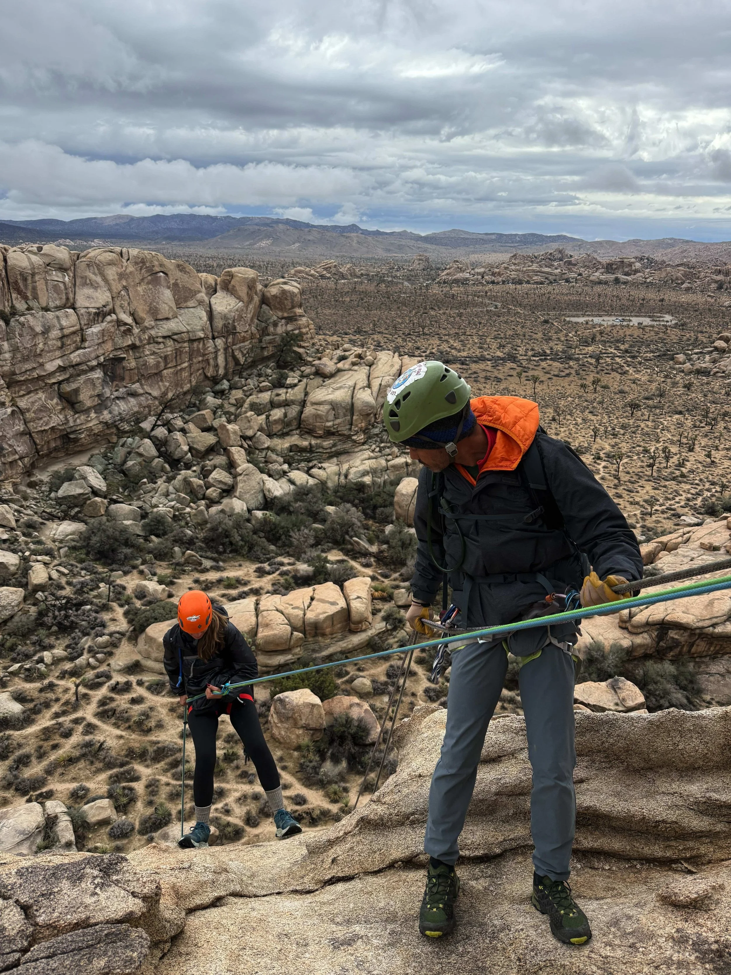 Two rock climbers with helmets and harnesses ascending a rocky cliff in a desert landscape, under cloudy skies.