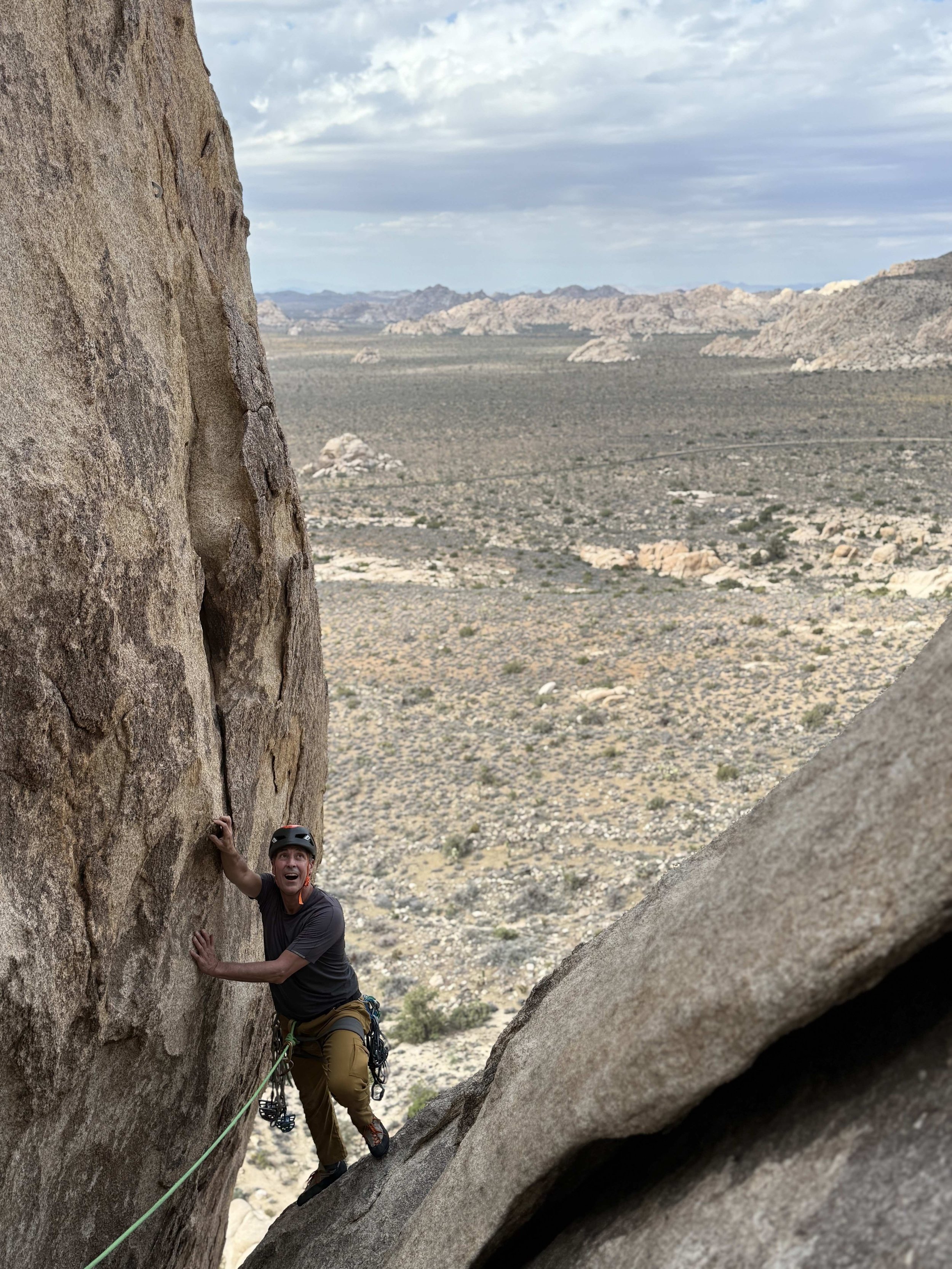 A person rock climbing on a steep rock face in a desert landscape, wearing a helmet and climbing gear, with a scenic view of mountains in the background.
