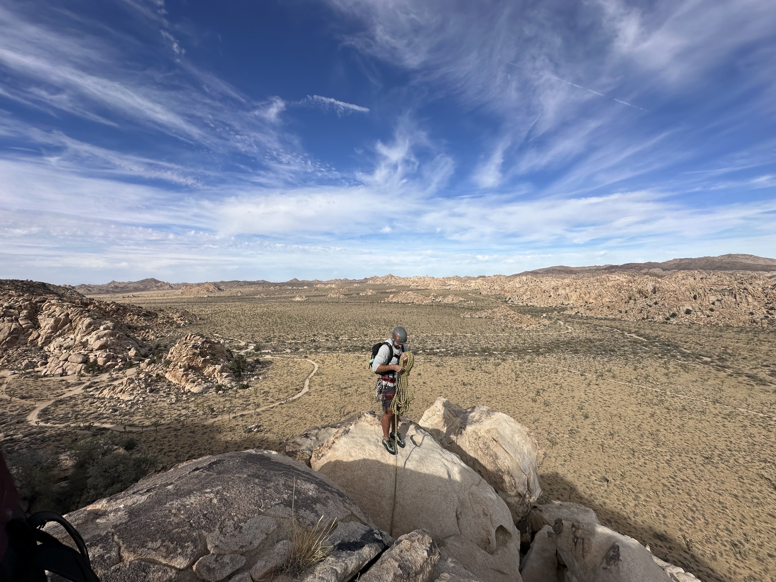 A person standing on a large rock in a desert landscape, holding a coiled rope, with distant mountains and a partly cloudy blue sky overhead.