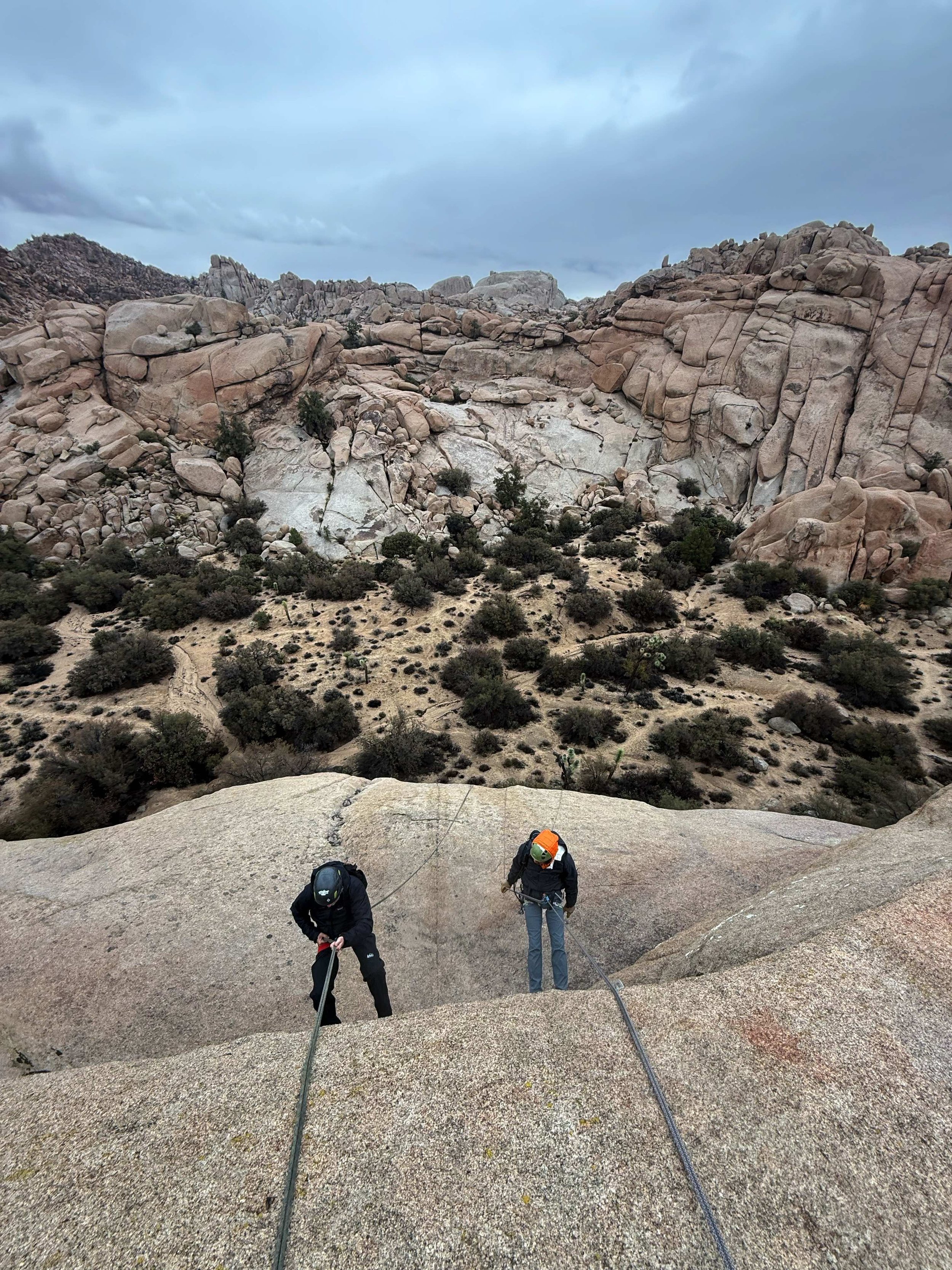 Two climbers ascending a steep rock face in a desert landscape with large granite boulders and sparse vegetation under a cloudy sky.