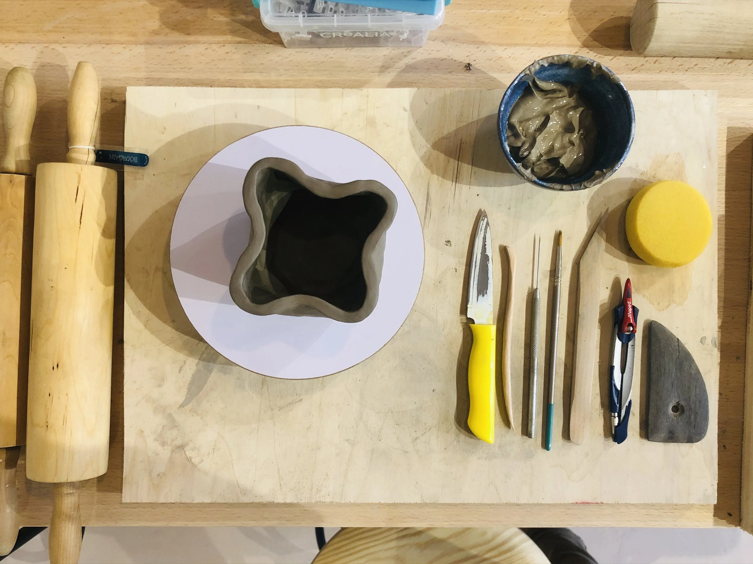 Table de travail avec outils de sculpture en céramique, une pièce en argile en cours de fabrication, pinceaux, cutters, éponge, une tasse de boue d'argile, et deux rouleaux