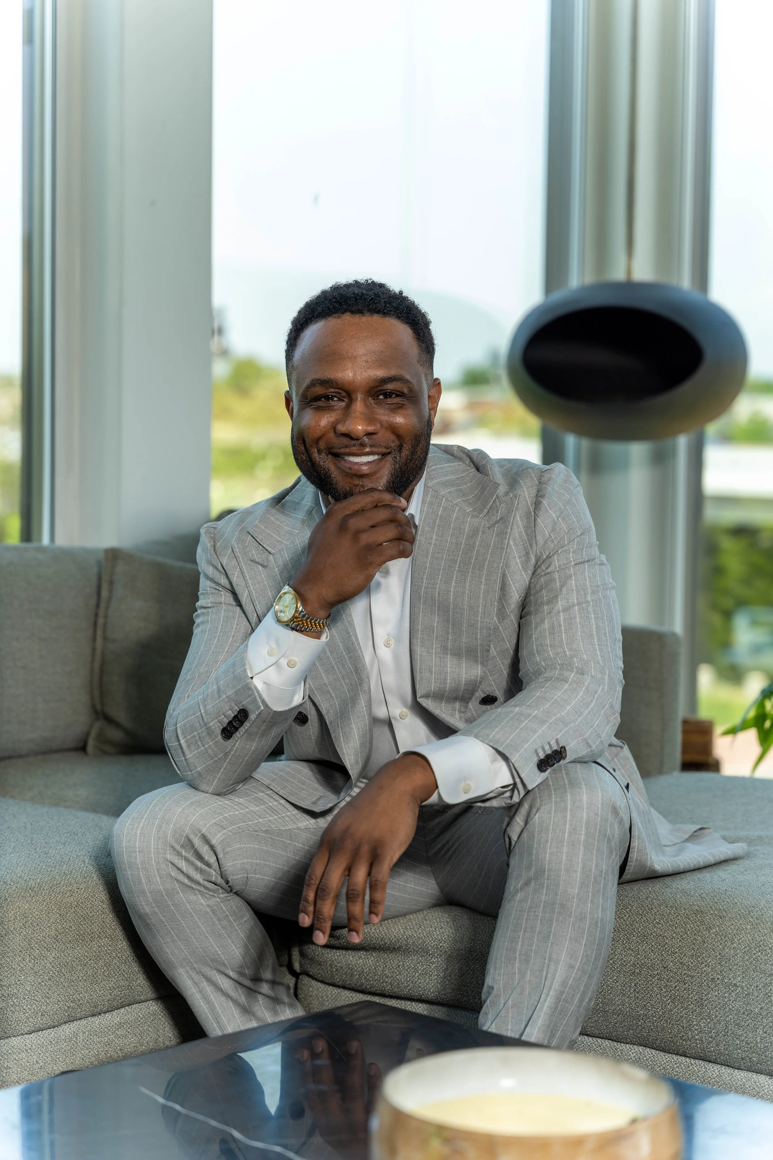 A smiling man in a light gray plaid suit sitting on a sofa in a modern indoor space, with a large window and greenery outside, while wearing a watch and a white shirt.