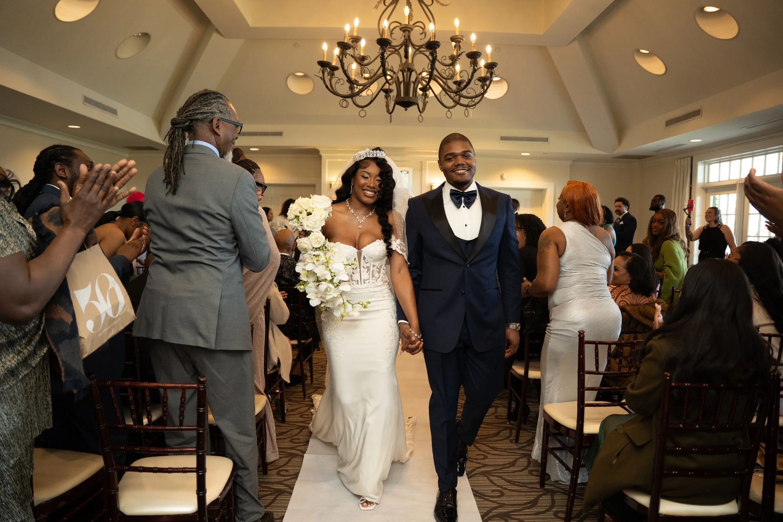 A bride and groom walk hand in hand down the aisle at their wedding ceremony, surrounded by guests who are clapping and smiling in a well-lit, elegant room with a chandelier hanging from the ceiling.