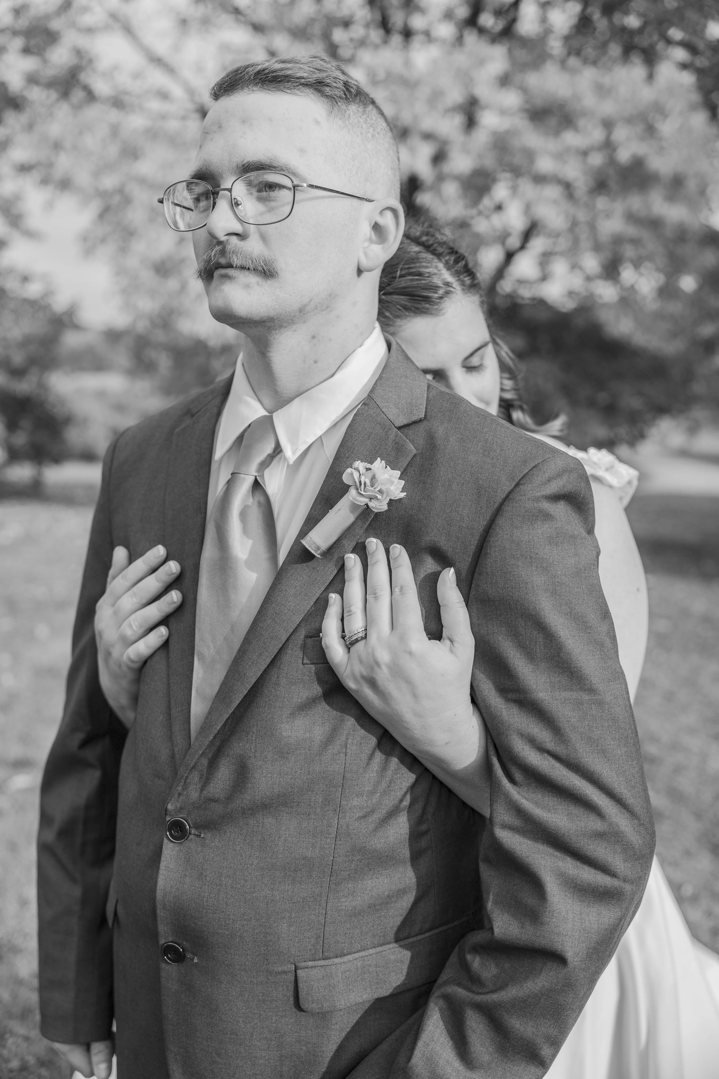 A black and white photograph of a groom in a suit with a boutonniere, standing outdoors with a woman behind him, who has her eyes closed and her hands on his chest, against a background of blurred trees.