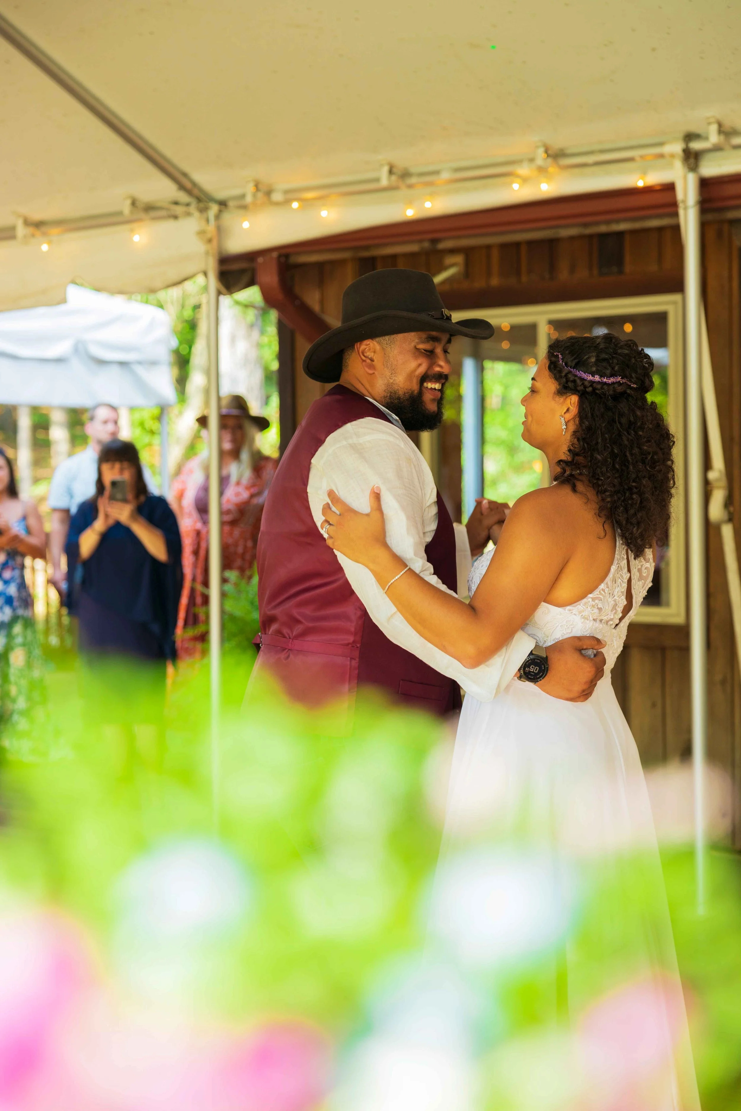 A man and woman are dancing closely together at an outdoor wedding reception, looking at each other and smiling. The man is wearing a cowboy hat and a vest, and the woman is in a white dress with curly hair and earrings. In the background, there are 