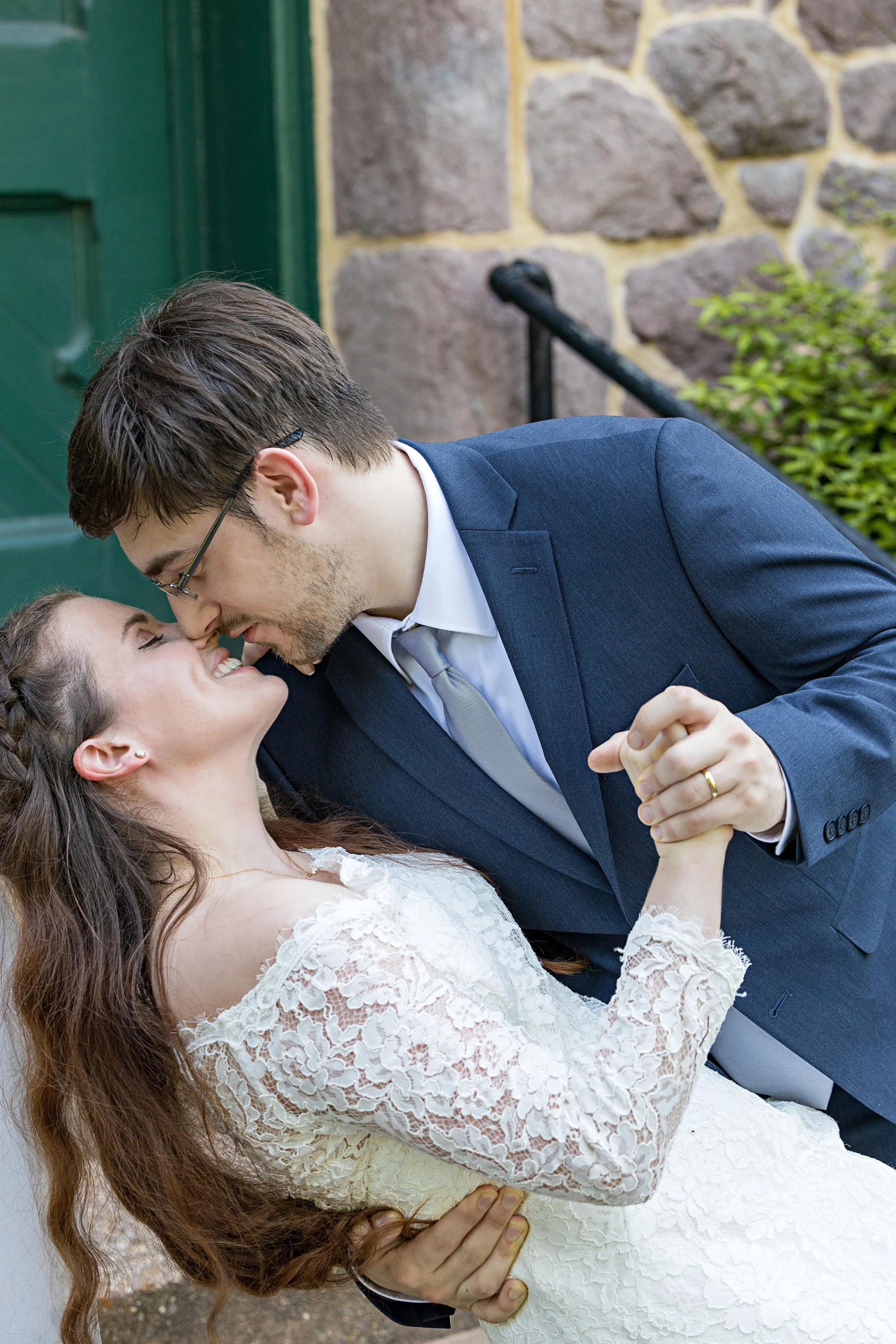A young couple, dressed in wedding attire, sharing a joyful moment outdoors with their foreheads touching, smiling, and singing. The bride has long, wavy brown hair and a lace wedding dress, and the groom has short brown hair, glasses, and a blue sui