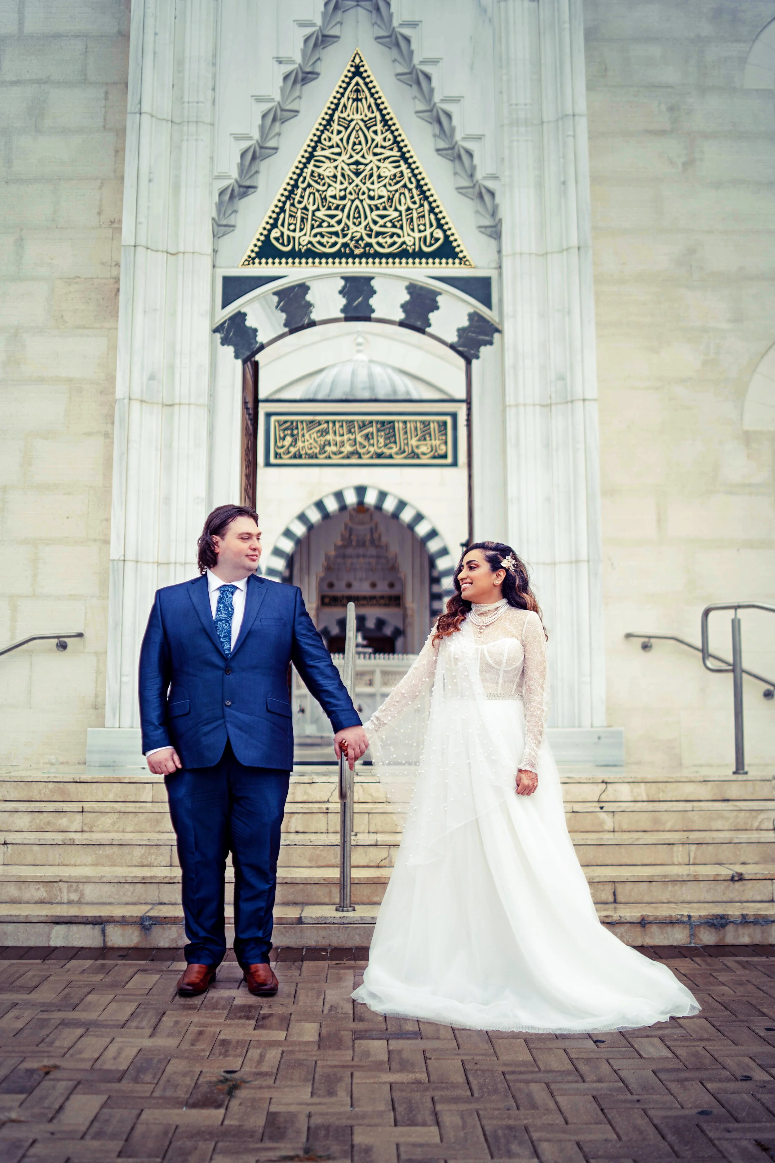 A bride and groom holding hands and standing on the steps of a building with Arabic calligraphy and geometric designs above. The groom is wearing a dark blue suit with a patterned tie, and the bride is wearing a white wedding gown with lace details. 