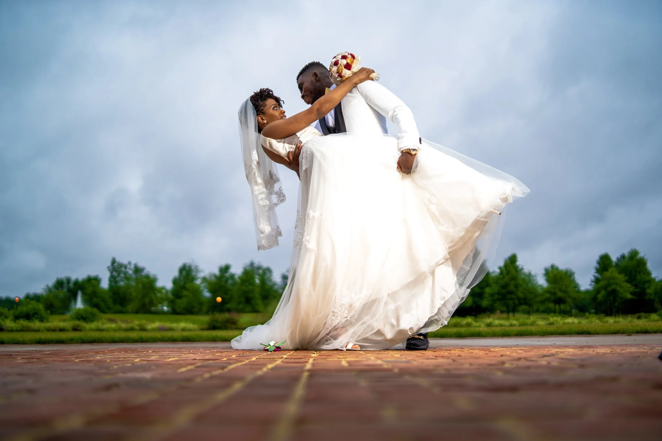 A bride and groom during a wedding ceremony outdoors, with the groom carrying the bride. The bride wears a white wedding dress and veil, while the groom is dressed in a white suit with a boutonniere. They are on a brick path with trees in the backgro