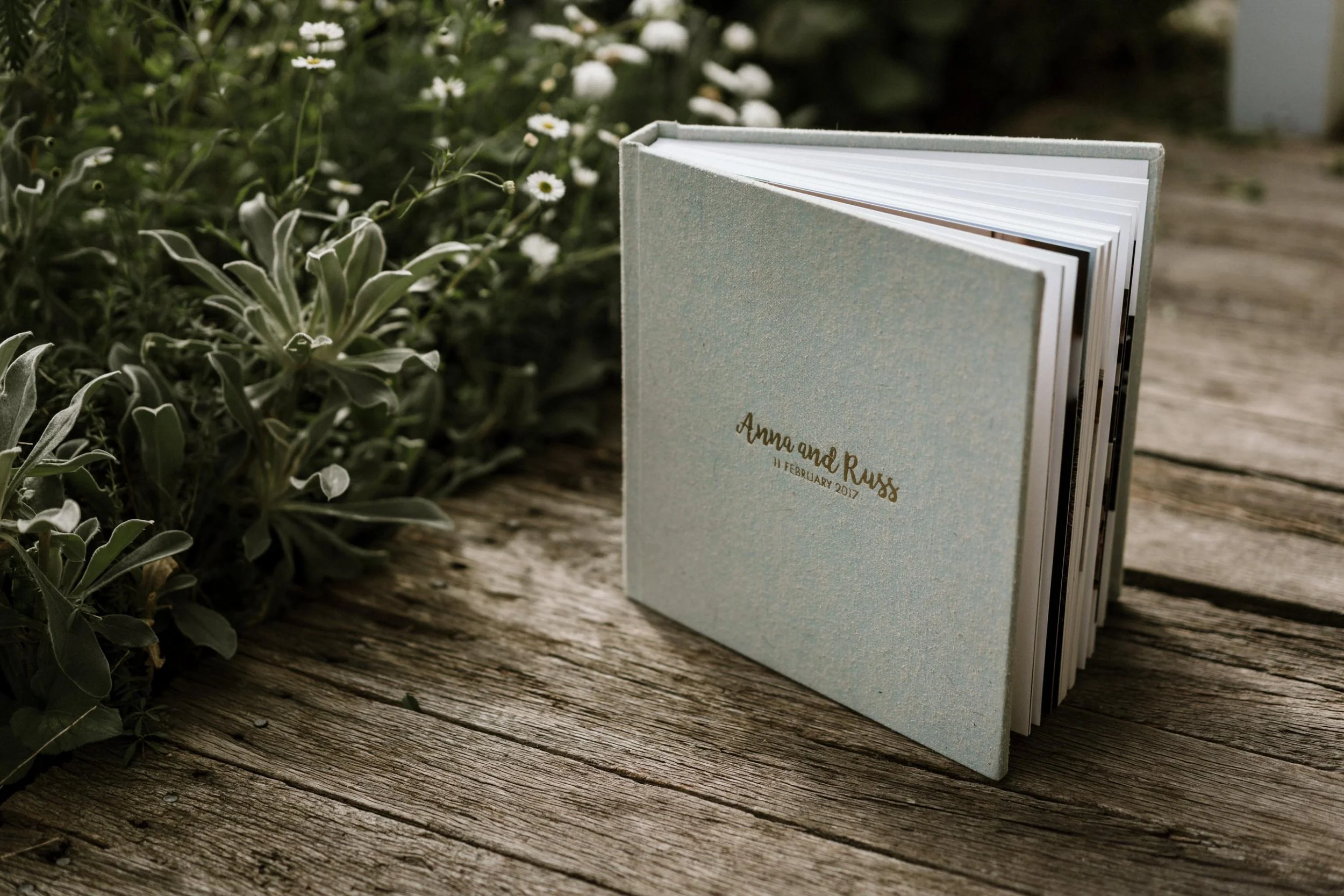 A wedding guestbook with a light blue cover facing open on a weathered wooden surface, with small white flowers and greenery nearby.