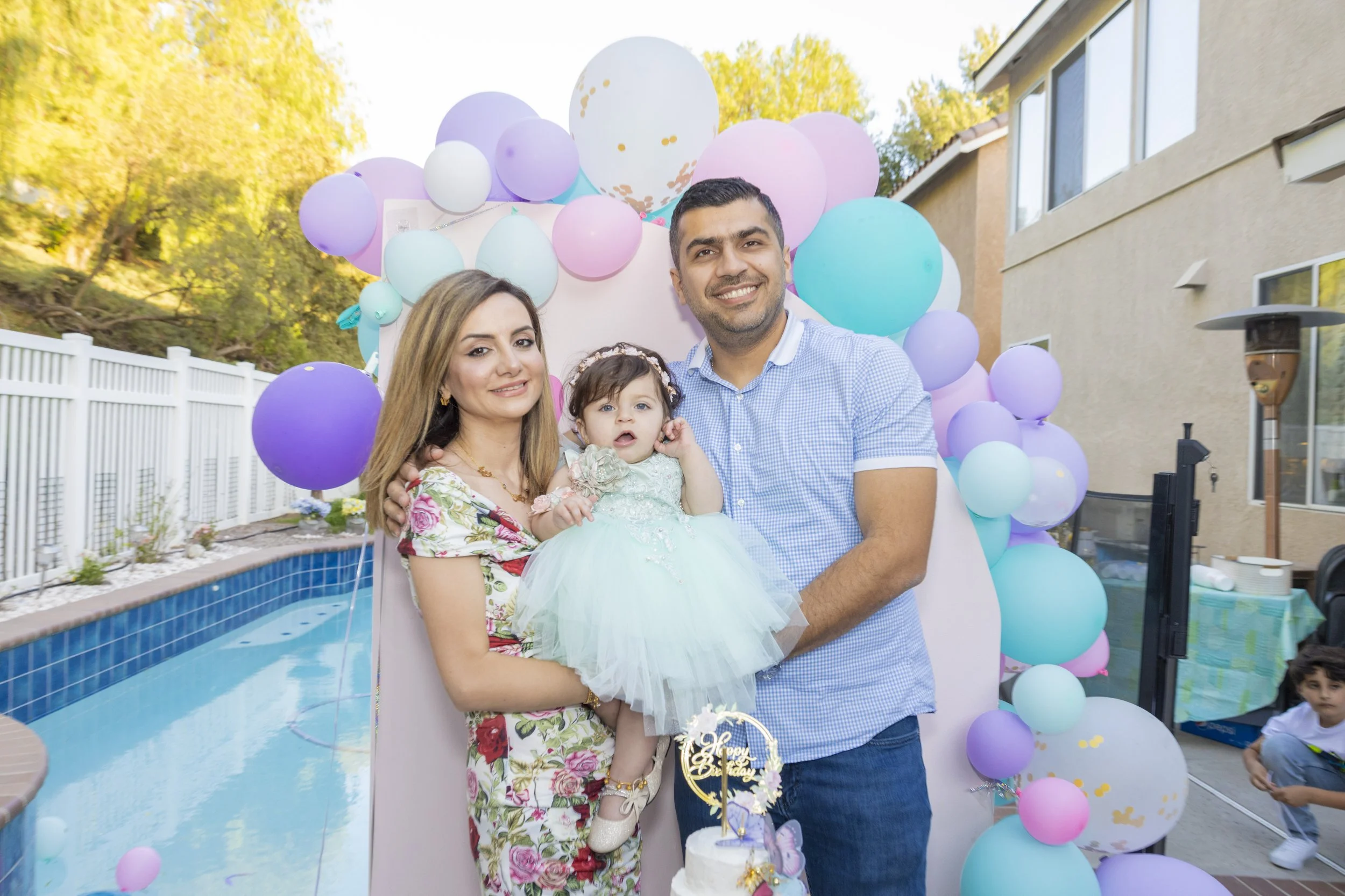 Family celebrating a birthday outdoors near a pool with purple, pink, and white balloons, with a house and trees in the background.
