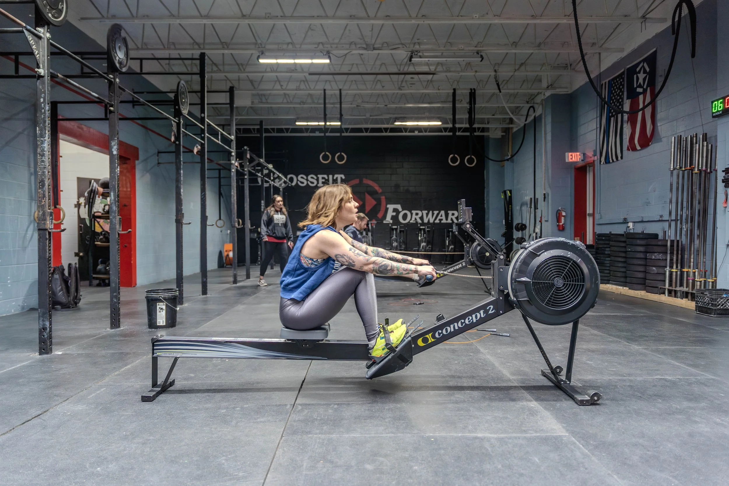 A woman with tattoos is seated on a rowing machine in a gym, focused on exercising, with other gym members and workout equipment visible in the background.