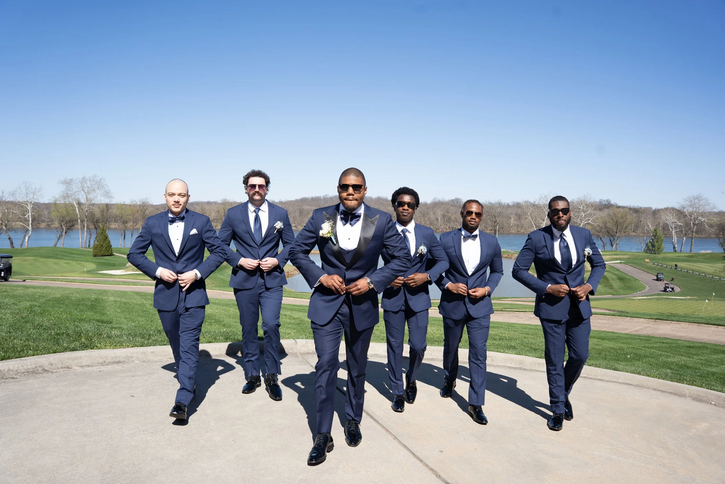 A group of seven men dressed in dark blue suits, white shirts, and black shoes walking on a paved path in a park with green grass, trees, and a lake in the background.