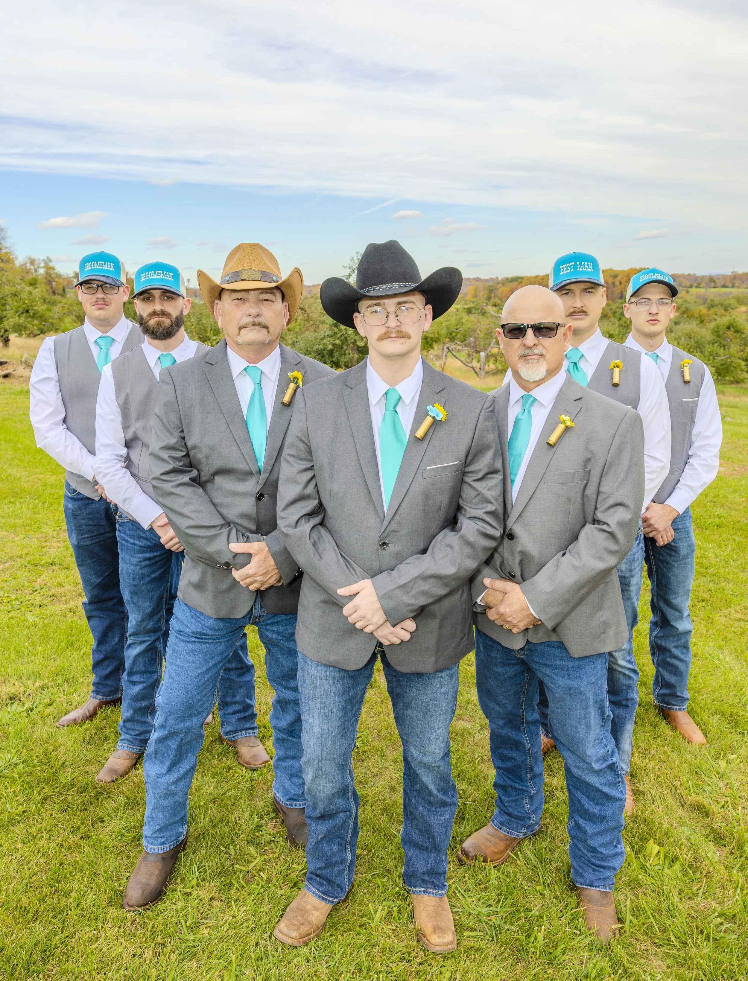 Six men dressed in formal suits and cowboy hats standing outdoors in a grassy field with trees and blue sky in the background.