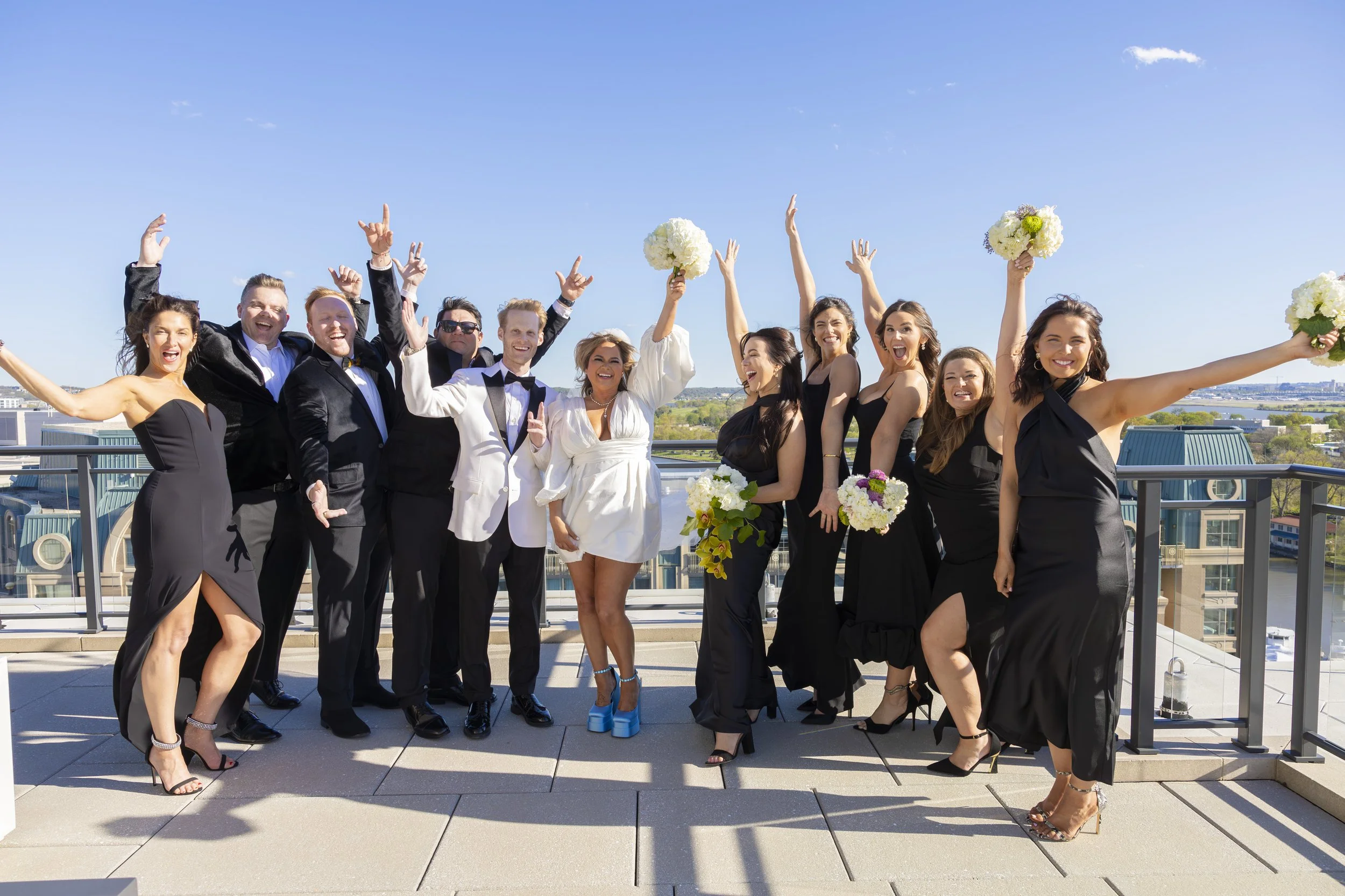 Group of wedding guests celebrating on a rooftop terrace with clear blue sky, some holding flowers, dressed in formal attire.