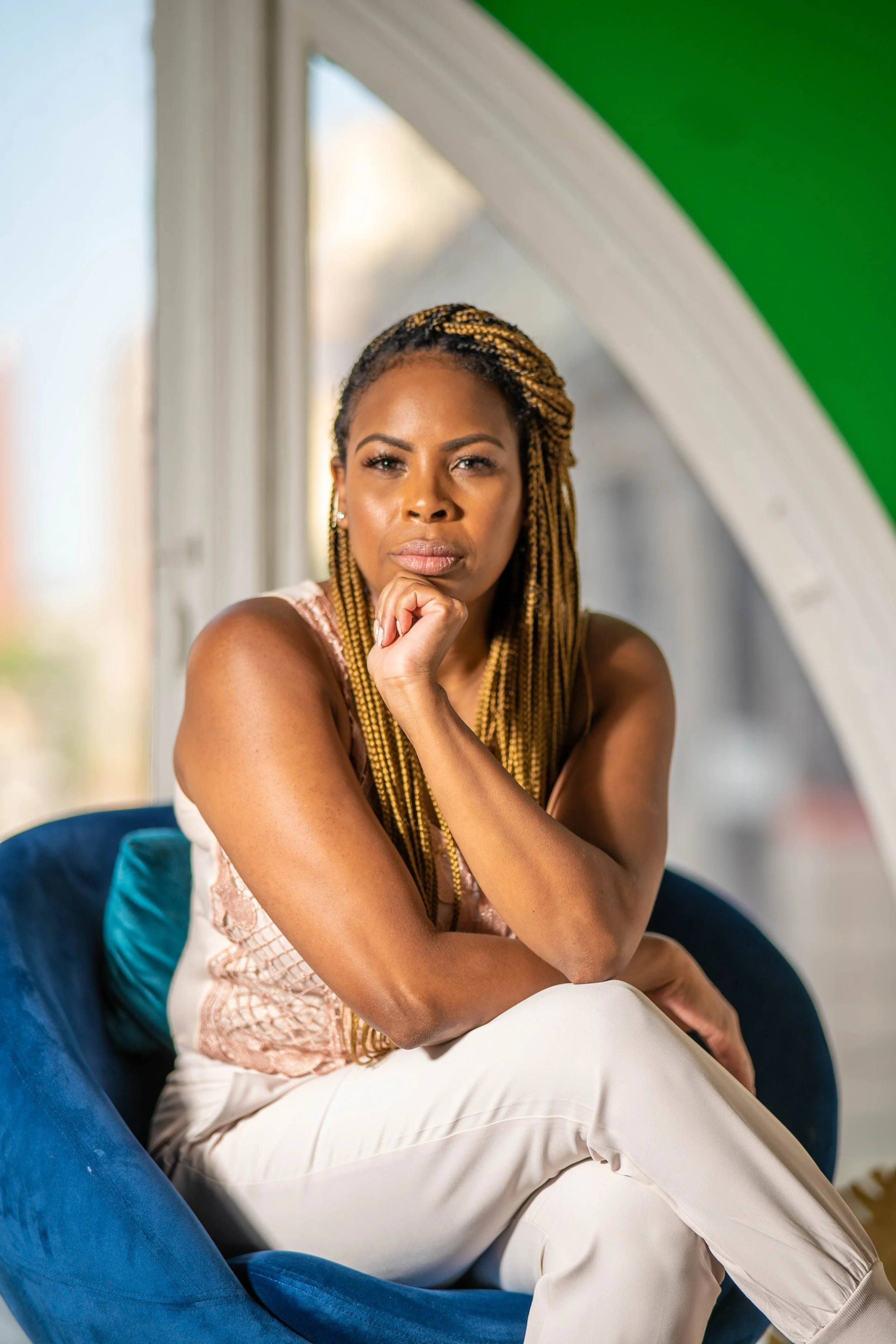 Portrait of a woman with braided hair, wearing a sleeveless top and white pants, sitting on a blue chair with a thoughtful expression, in a room with window and green-painted ceiling.
