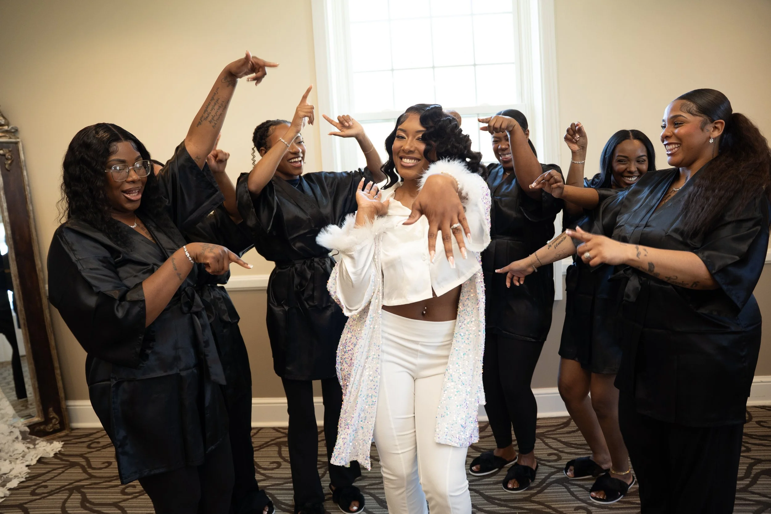 A bride in white pants and a jewel adorned jacket poses with her bridal party, who are wearing black satin robes, in a well-lit room with beige walls and a patterned carpet.