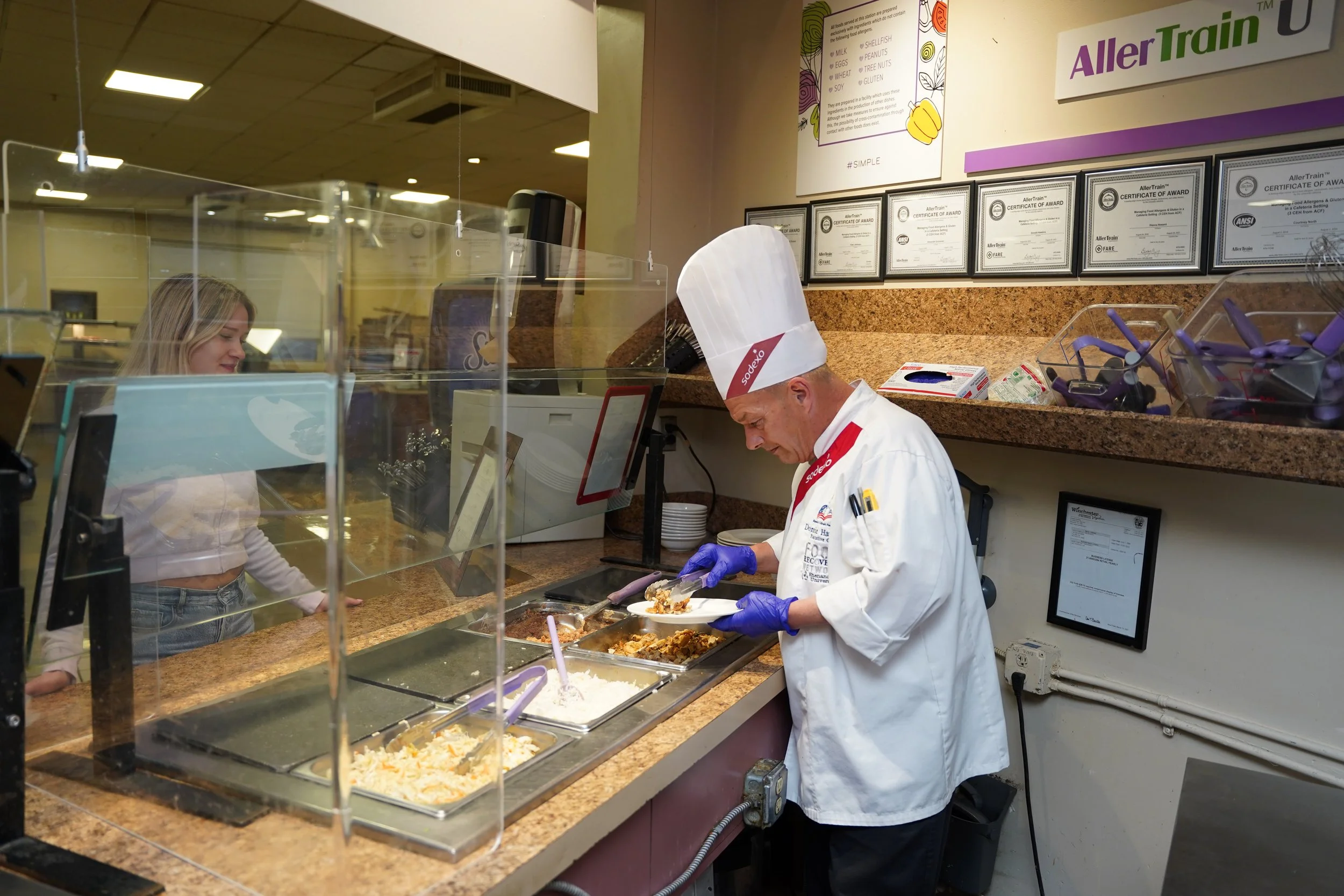 A woman in a white top and jeans stands behind a clear safety partition at a cafeteria serving counter. An older man wearing a chef's hat, white coat, and purple gloves is serving food onto a plate. The counter has trays of food, including mashed pot