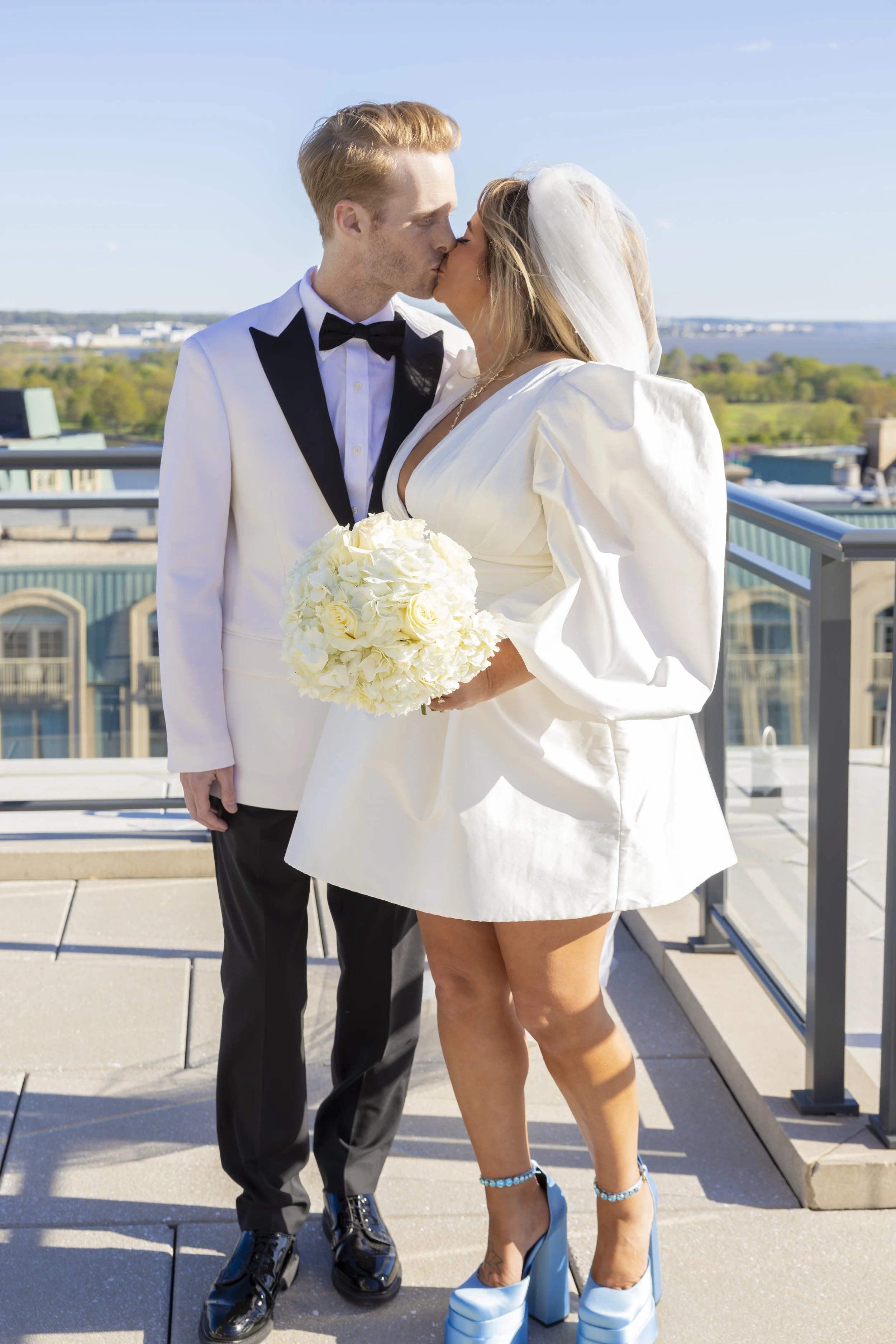 A couple dressed in wedding attire sharing a kiss on a rooftop balcony with a city and water in the background.