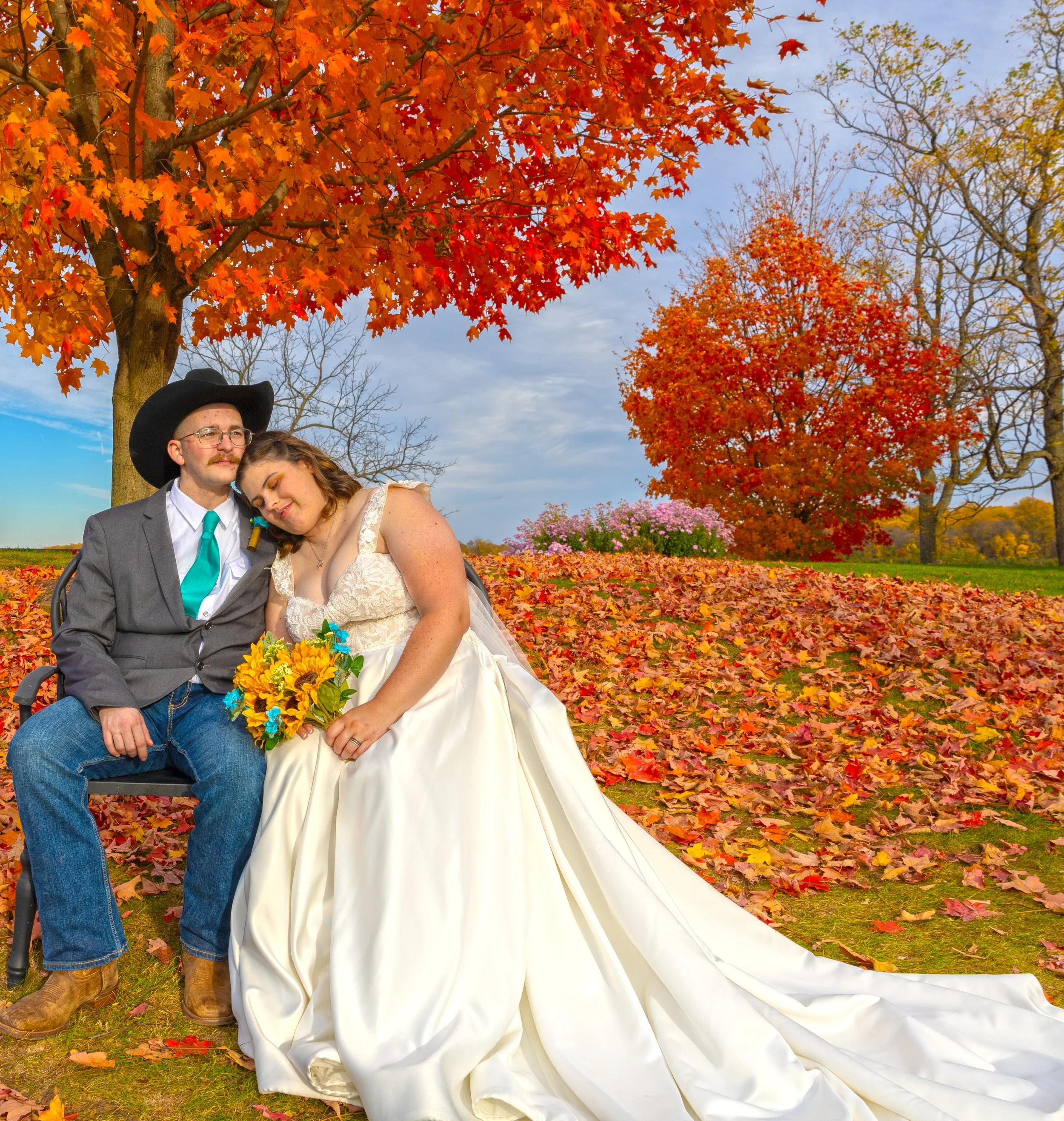 A couple sitting outdoors during fall, with autumn trees and colorful fallen leaves surrounding them.