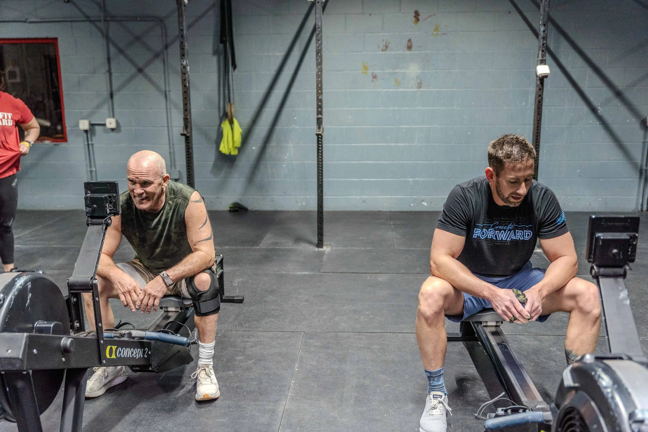 Two men seated on workout benches in a gym, resting after exercise, with gym equipment and a gray wall in the background.