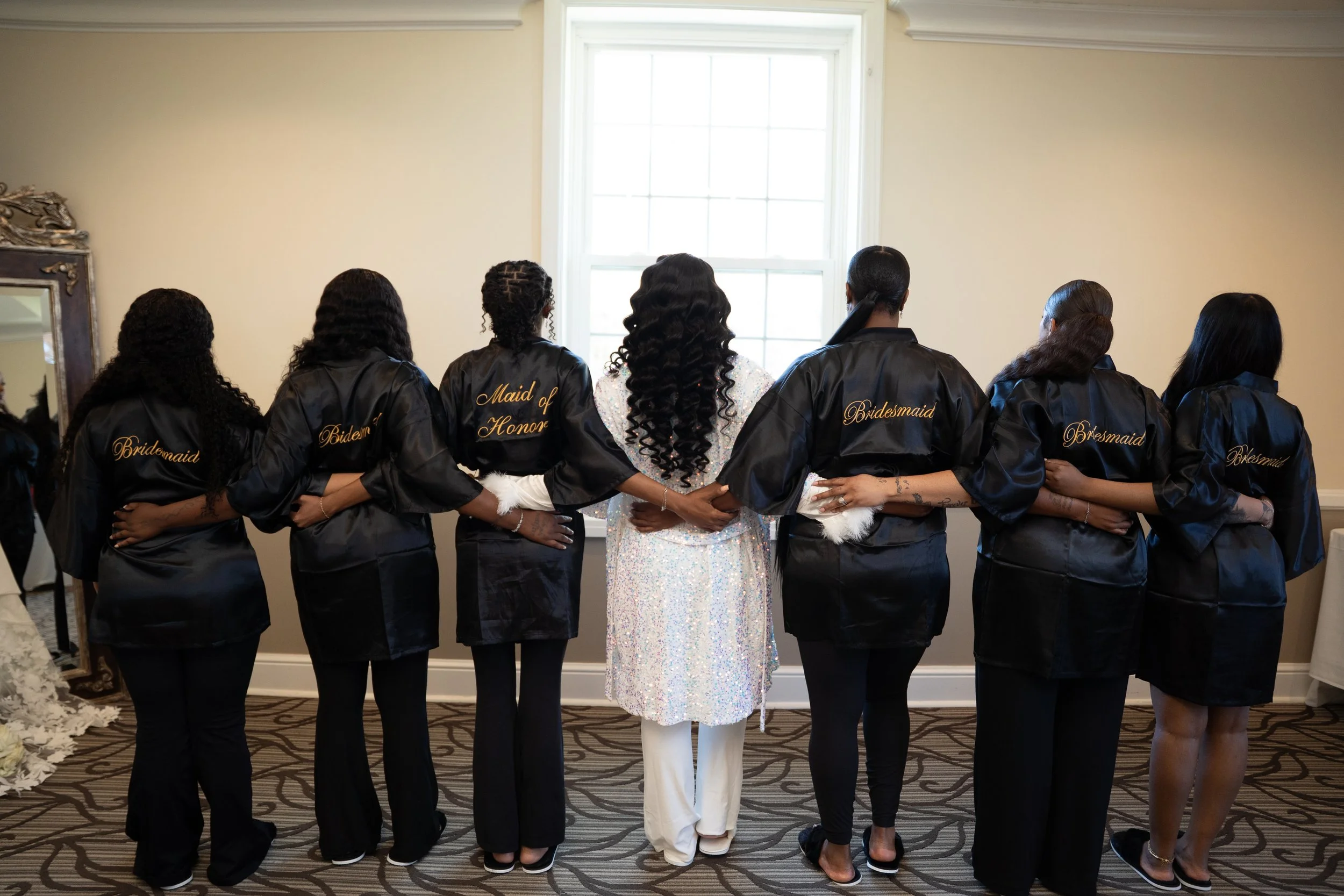 A bride and her six bridesmaids standing together in a room, facing away from the camera. The bridesmaids are wearing black satin robes with 'Bridesmaid' embroidered on the back, and one has 'Maid of Honor'. The bride is in the center, wearing a whit