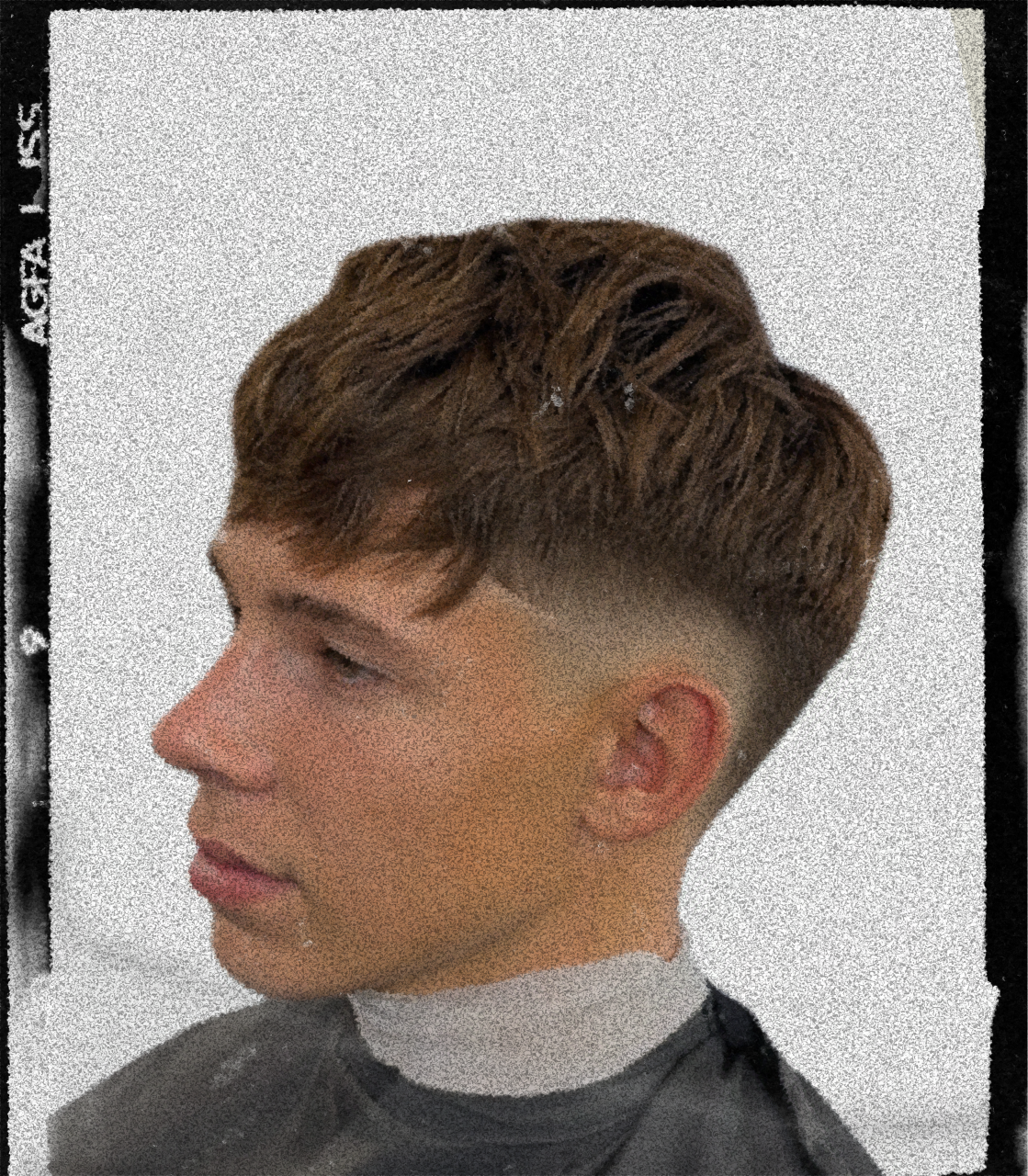 Side profile of a young man with a textured fade haircut, wearing a black apron and a white shirt, against a plain wall background.