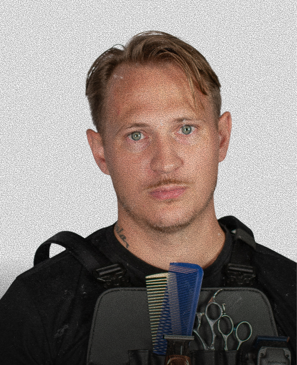 A young man with light brown hair, green eyes, and a serious expression, wearing a black uniform with a tactical vest, holding a hairdressing comb and scissors.