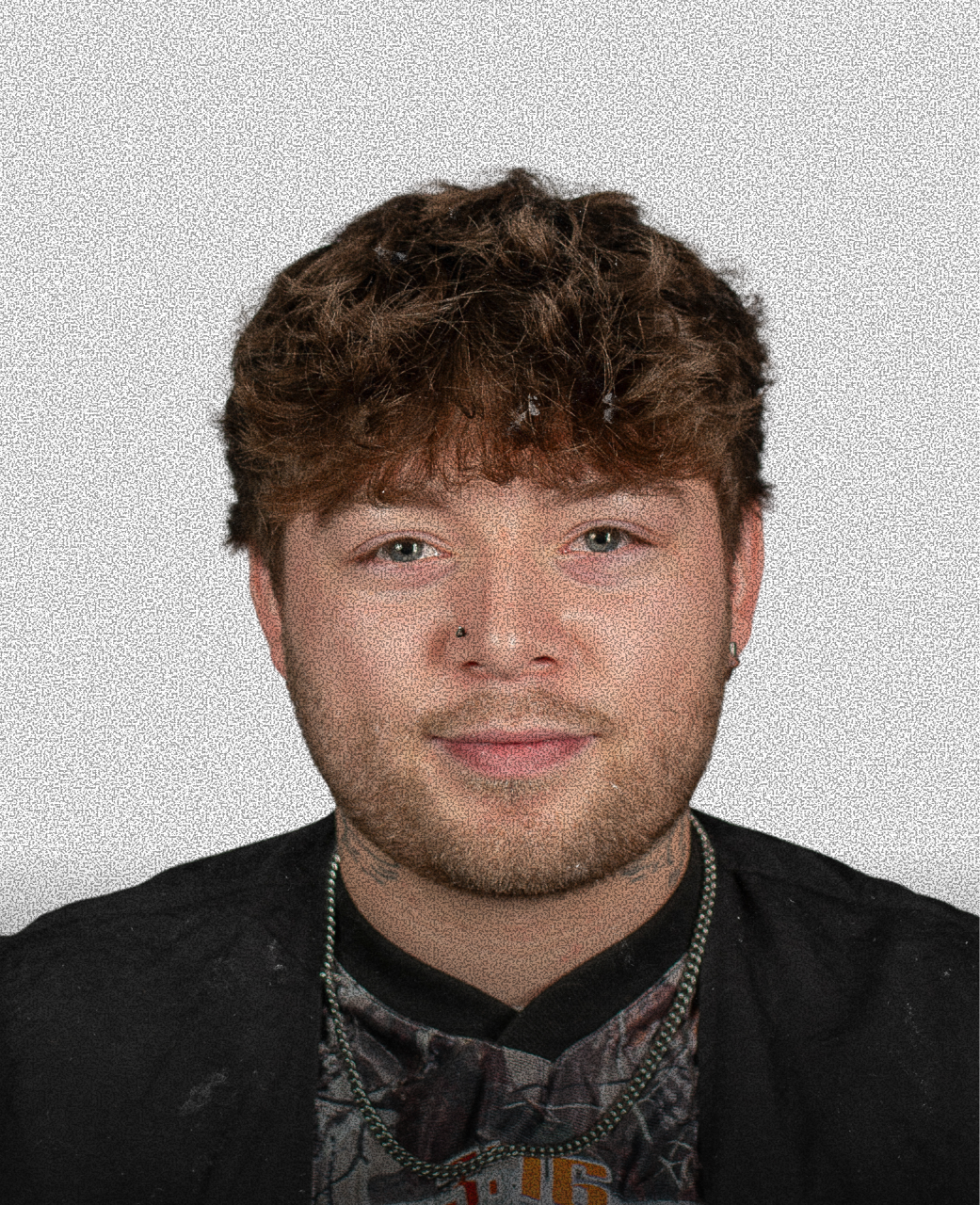 A young man with curly brown hair, wearing a gray and black patterned shirt, a black jacket, and multiple necklaces, with a light gray background.