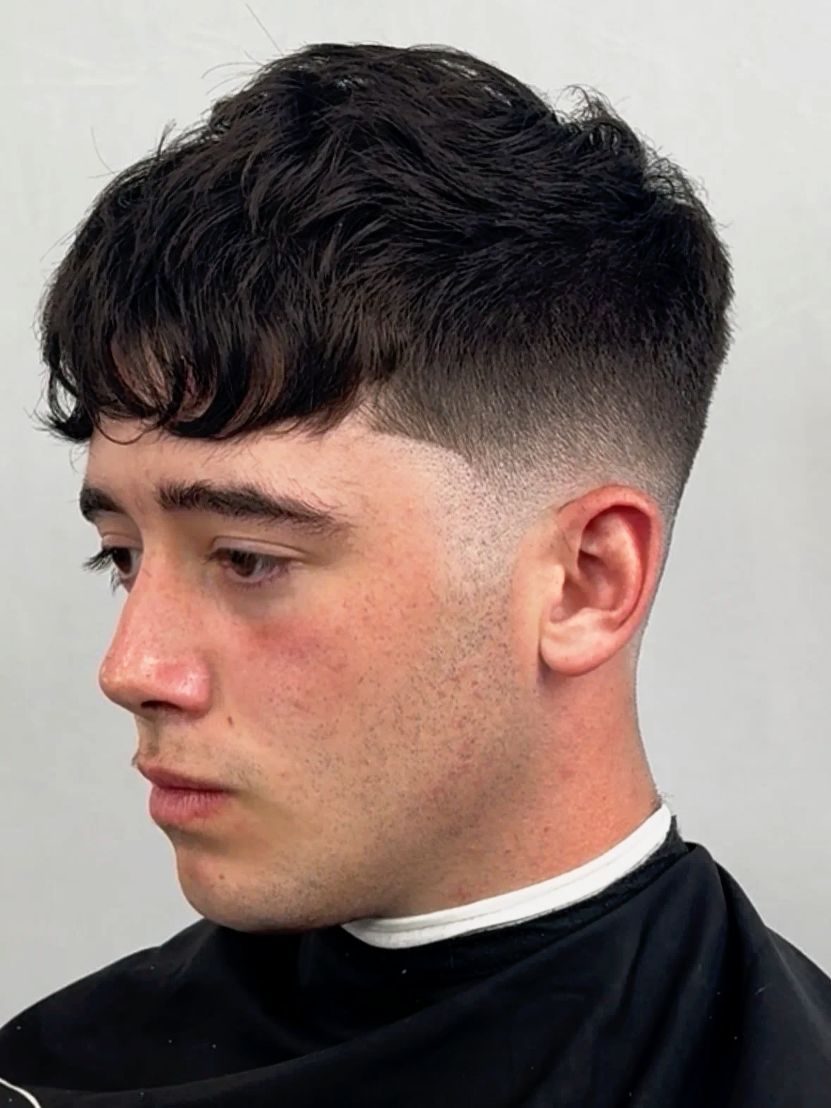 A young man with dark, wavy hair and a textured haircut, wearing a black barber cape, looking to the side in a barbershop.