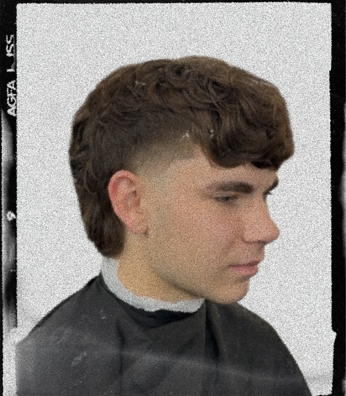 Side profile of a young man with styled brown hair, wearing a black barber cape and a white turtleneck underneath, in a hair salon.