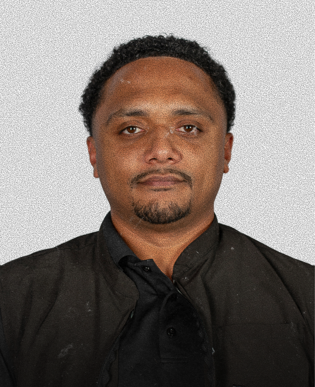 A man with dark curly hair, facial hair, and a serious expression, wearing a black shirt against a plain light-colored background.
