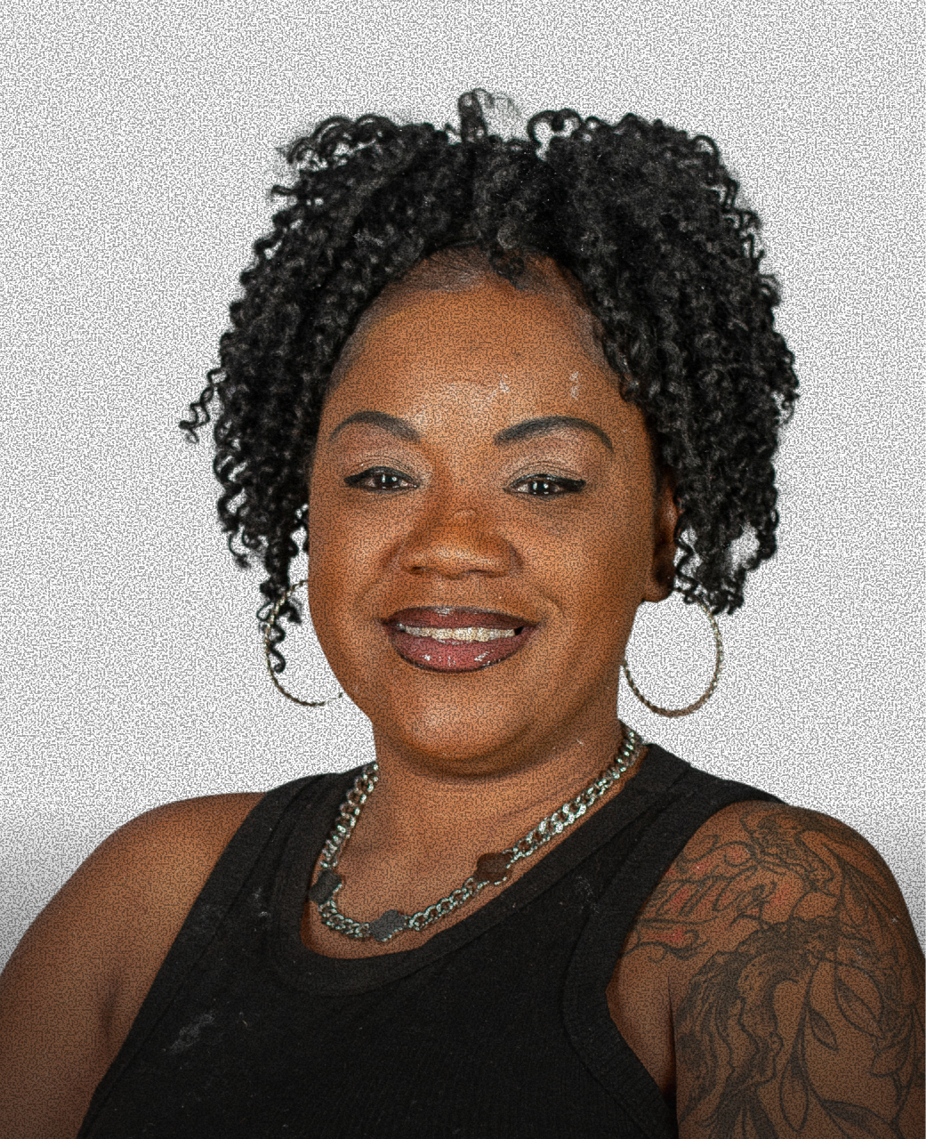 A woman with curly black hair and hoop earrings, smiling, wearing a black sleeveless top, and a necklace with large links, standing against a plain light background.