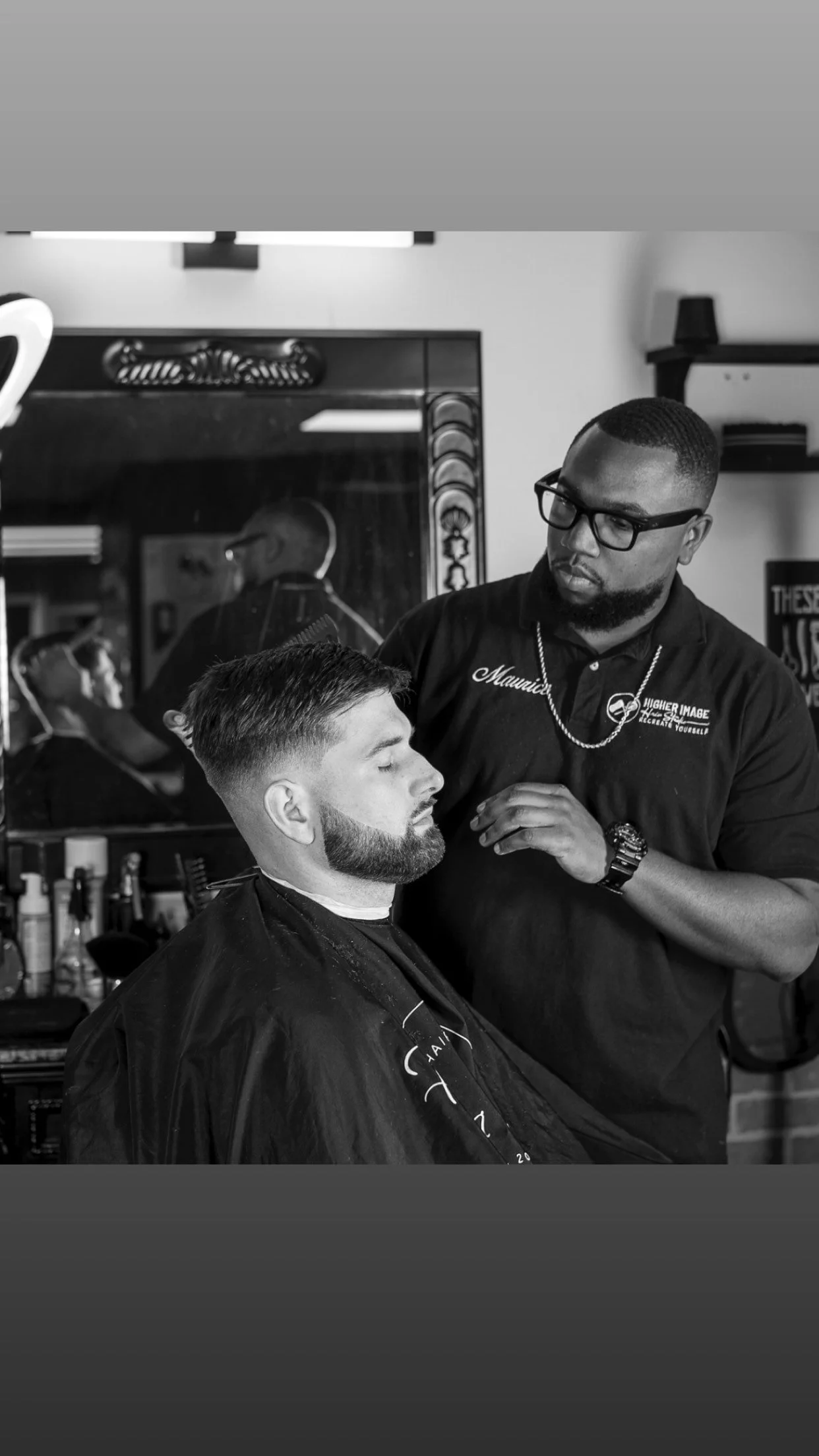 A man with a beard and short hair gets his hair cut at a barbershop by a barber wearing glasses and a chain, with tools and products visible on the counter behind them.