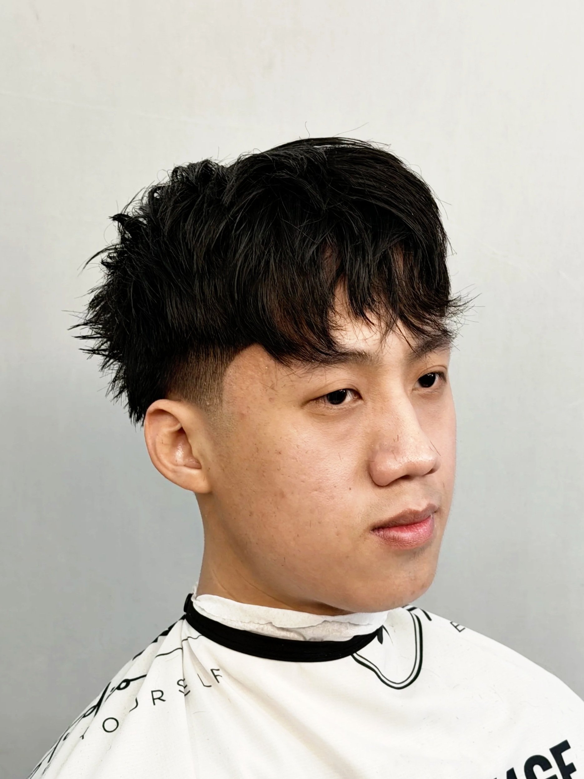 A young man with short, black, wavy hair and light skin, wearing a white haircut cape, looking to the side against a blurred background.