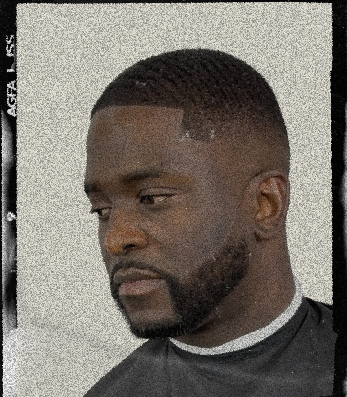 Close-up of a young man with a beard, short textured haircut, wearing a black shirt with white collar, looking to the left against a plain light-colored background.