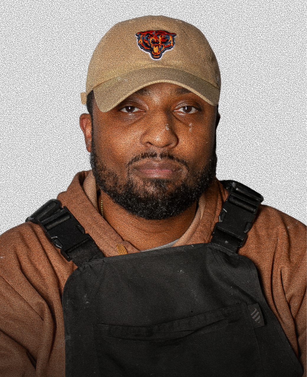 A man with a beard wearing a tan Chicago Bears cap, a brown shirt, and a black vest, against a light grey background.