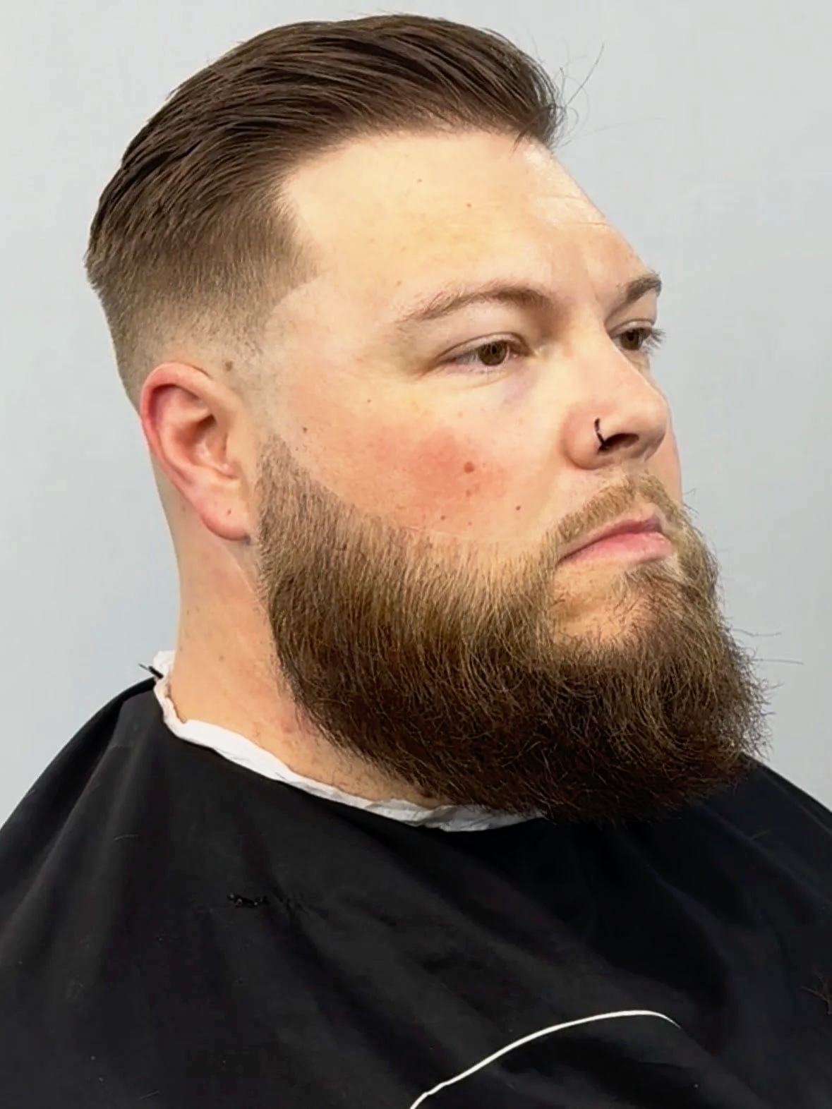 A man with styled brown hair and a full beard, wearing a black cape, at a hair salon, looking to his right.