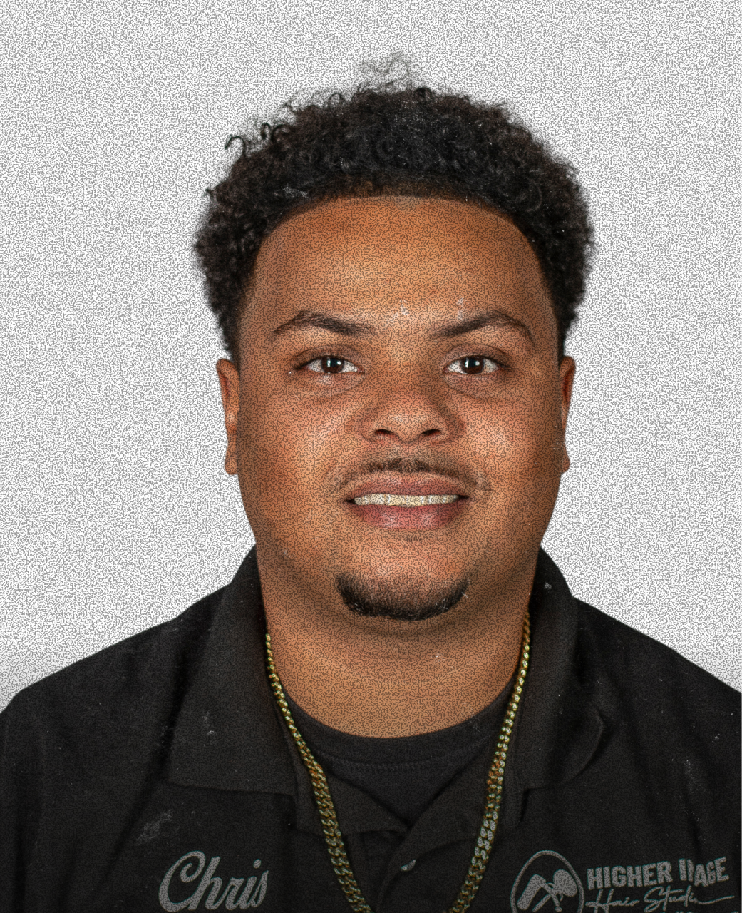Headshot of a young man with curly hair, wearing a black shirt with embroidery, and a gold chain necklace, smiling slightly against a plain background.
