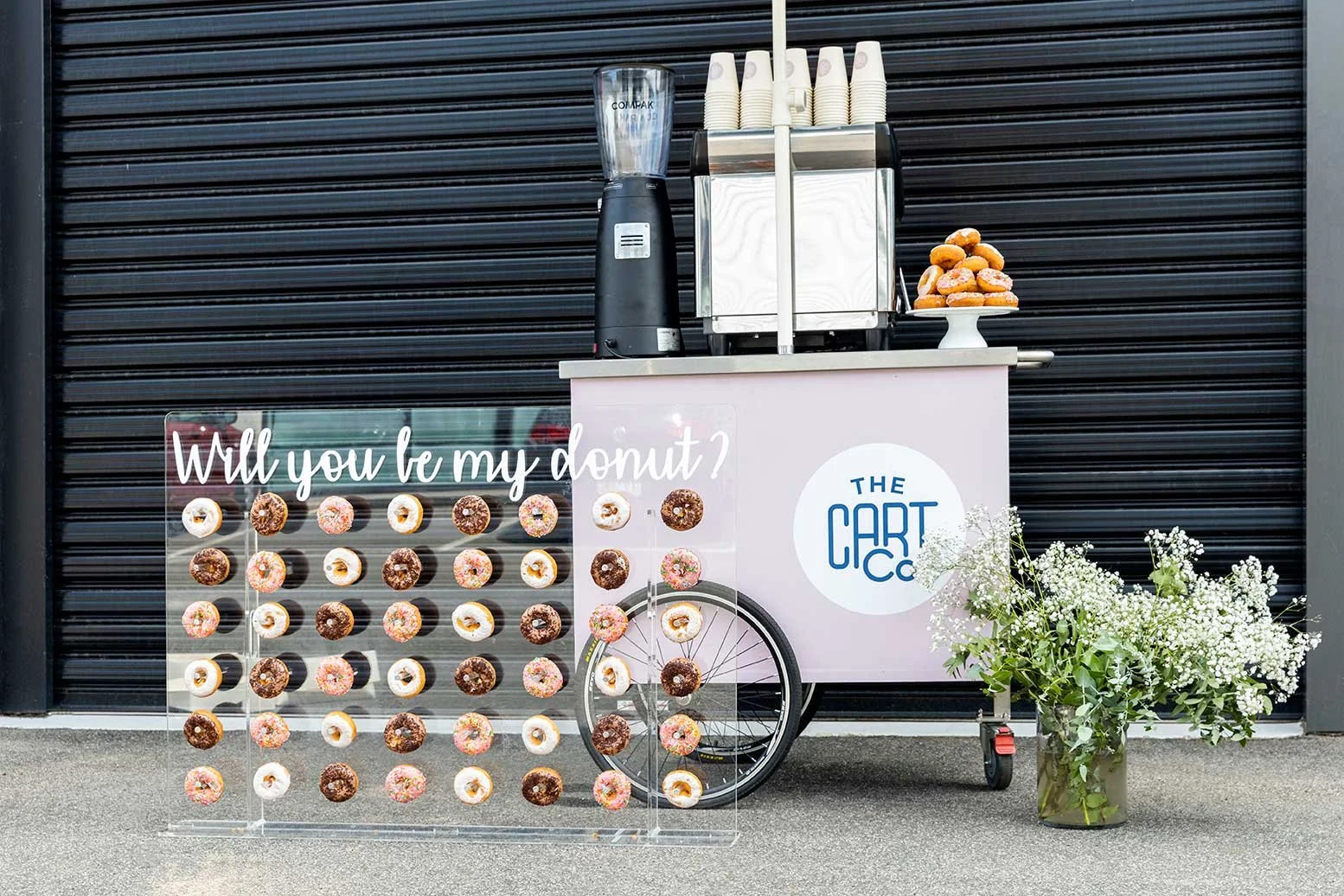 Donut wall displaying assorted donuts beside a pink The Cart Co coffee cart