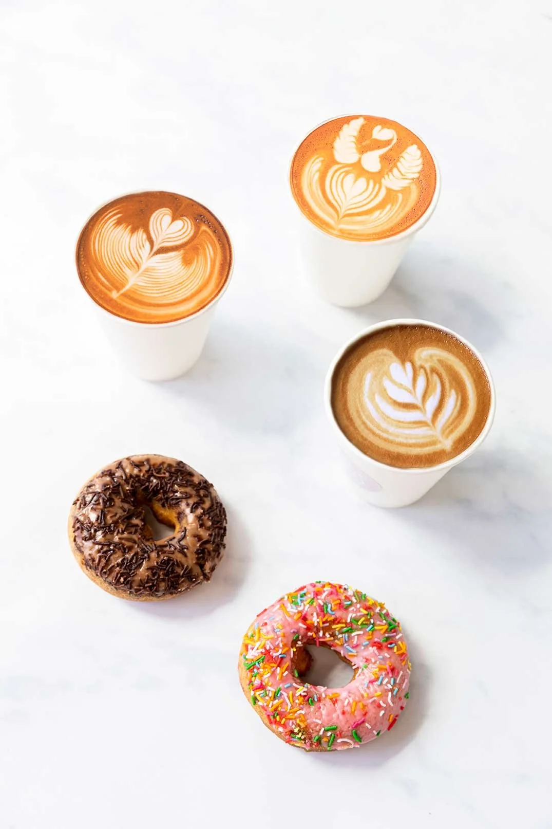 Three cups of coffee with latte art and two iced donuts arranged on a light surface