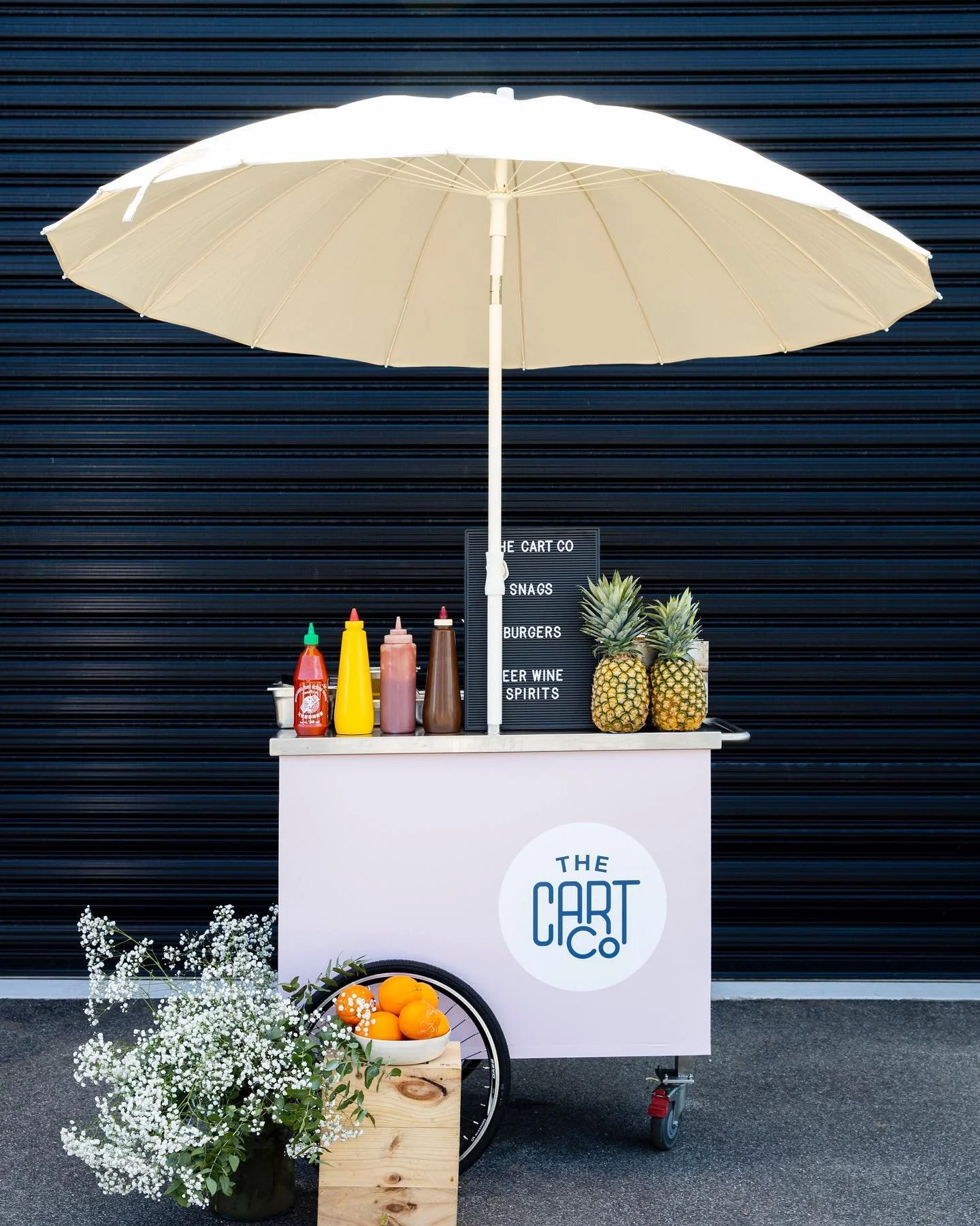White The Cart Co beverage cart with a large cream umbrella, bottled sauces and pineapples outdoors