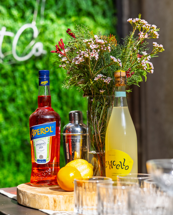 Aperol and limoncello bottles displayed with citrus fruit, cocktail glasses and flowers