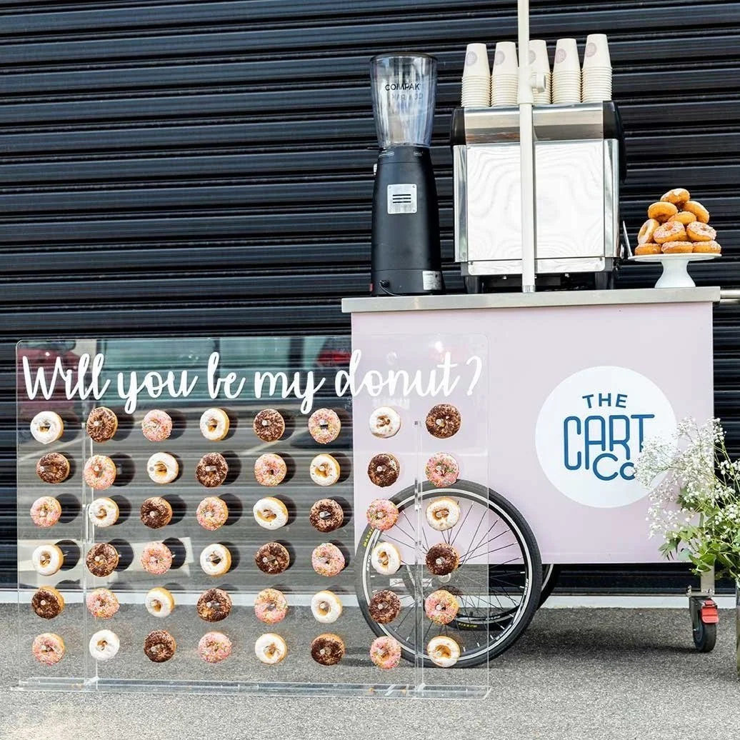 Donut wall displaying assorted donuts beside a pink The Cart Co coffee cart