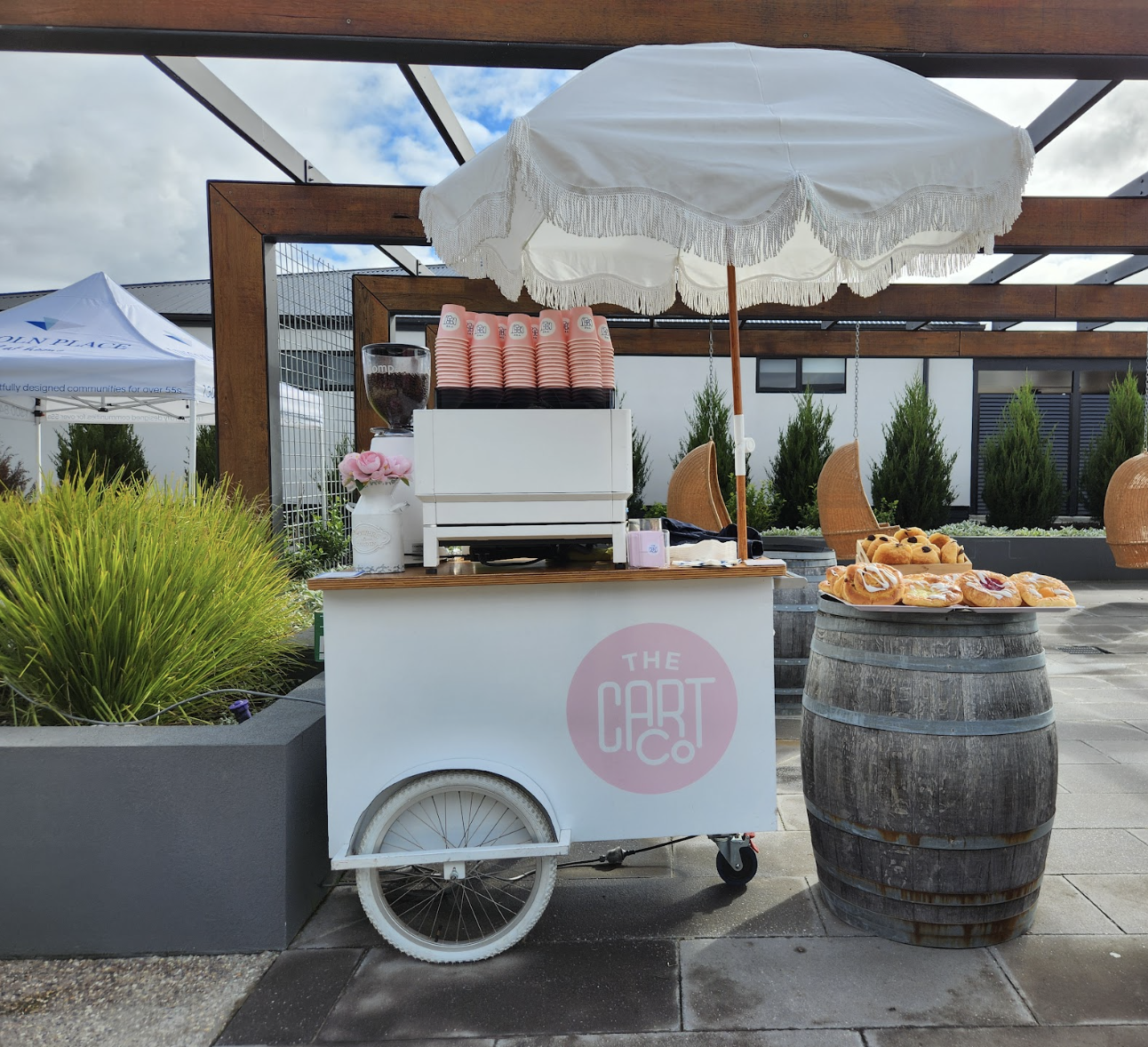 White coffee cart with umbrella and espresso machine beside a pastry display outdoors