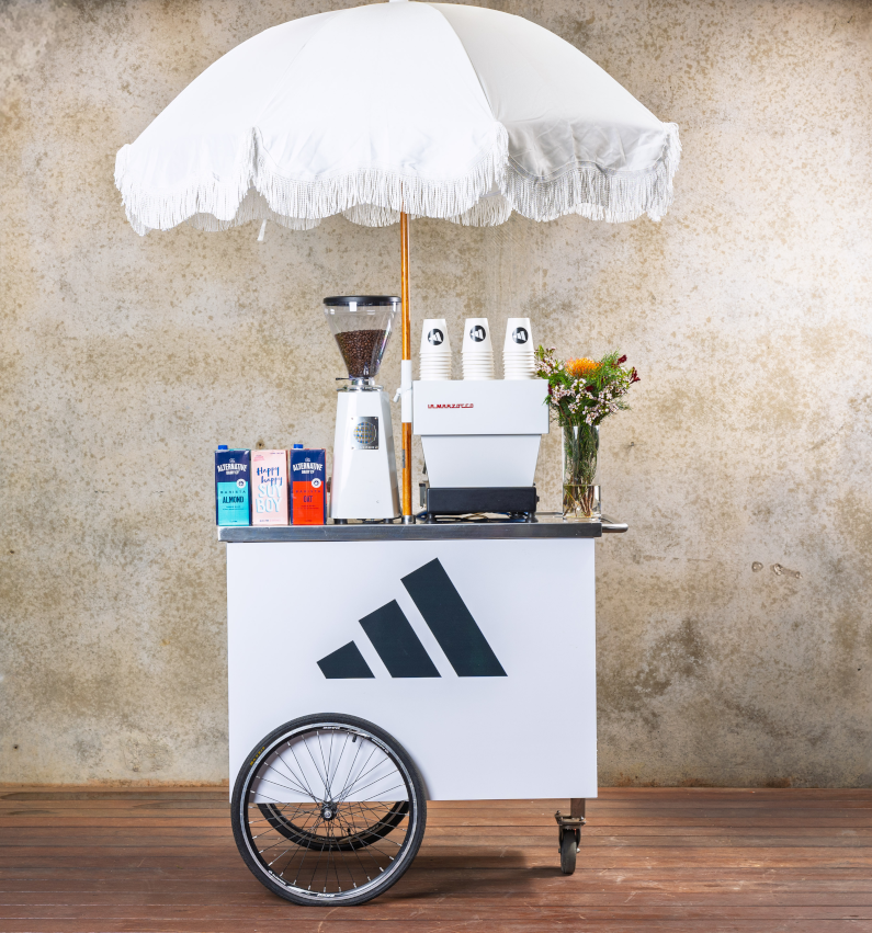 Adidas-branded coffee cart with white umbrella, espresso machine and coffee grinder