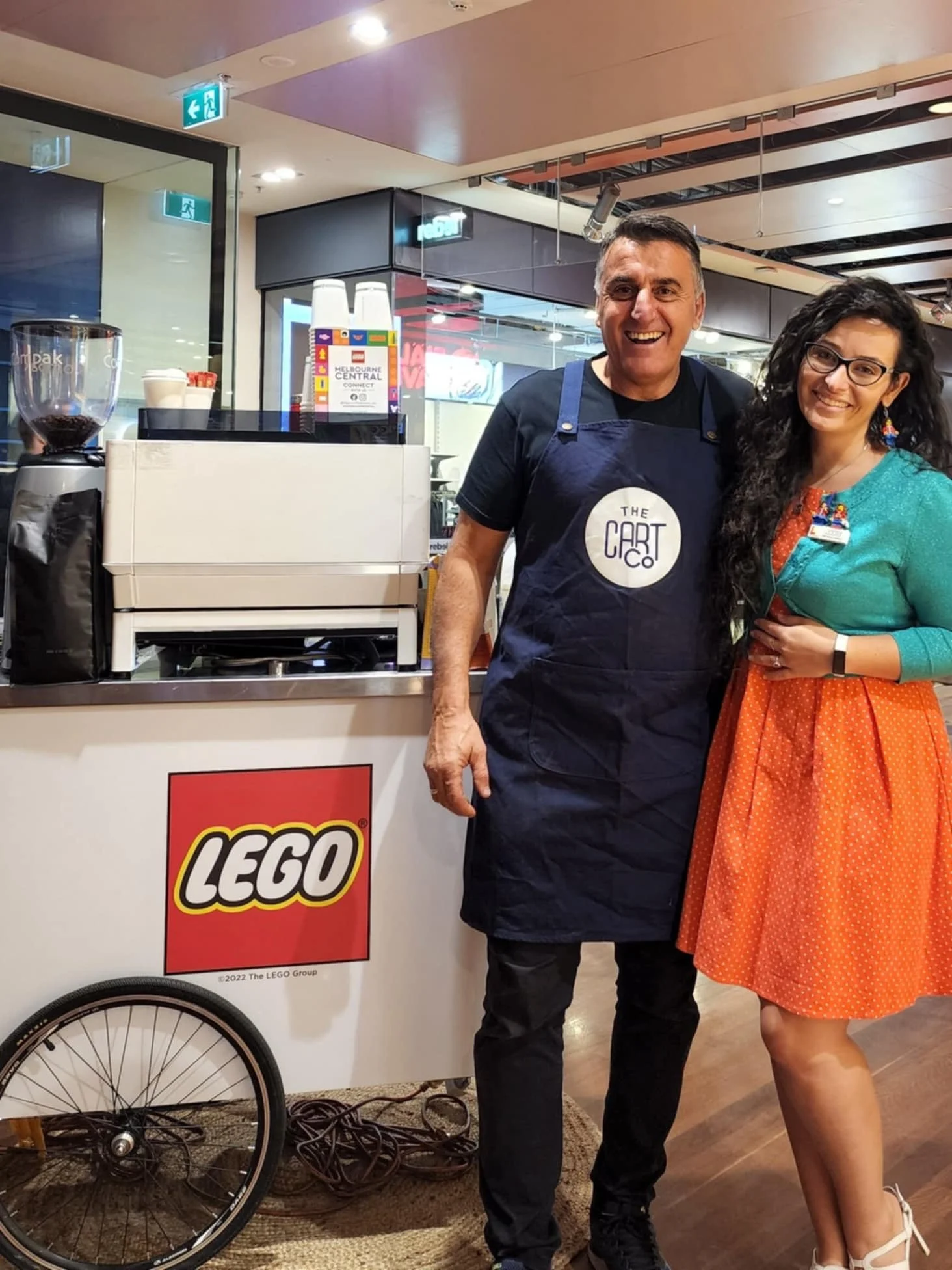 A barista and guest beside a LEGO-branded mobile coffee cart at an event